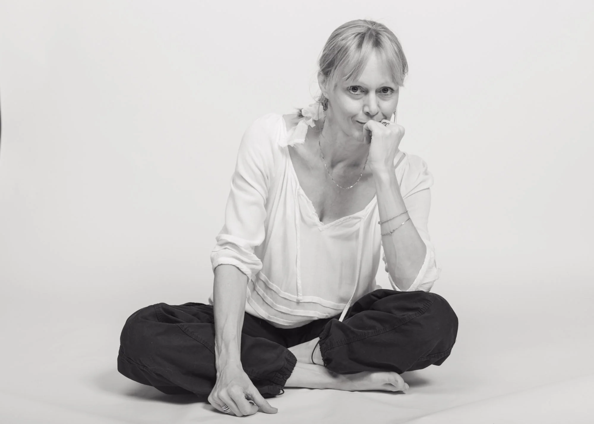 A woman sitting cross-legged on the floor, wearing a white top and dark pants, with her right hand resting on her knee and her left hand near her mouth, looking directly at the camera with a thoughtful expression.