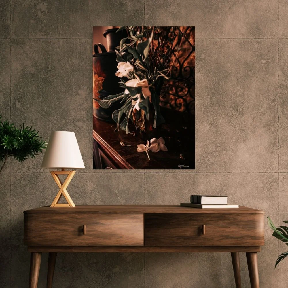 A wooden console table with a white table lamp, a few books, and a plant on the left. A framed floral still life painting hangs on a gray wall above the table.