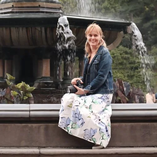 A woman in a denim jacket and floral skirt sitting on a fountain's edge holding a camera, with water flowing from the fountain behind her.