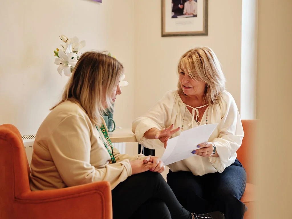 Two women having a discussion in an office or home setting. One woman holds papers, and the other listens attentively.