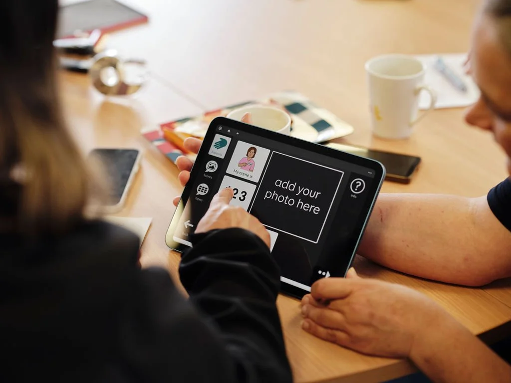 Two people sitting at a table with a tablet that has an app open, displaying a message to add a photo, surrounded by coffee mugs and phones.