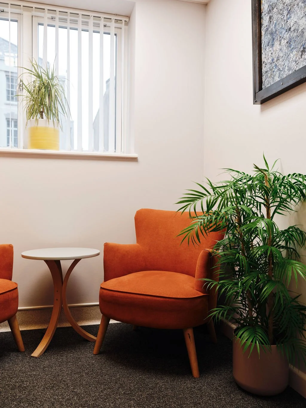 A cozy corner with an orange armchair, a small round wooden table, and green potted plants near a window with vertical blinds.