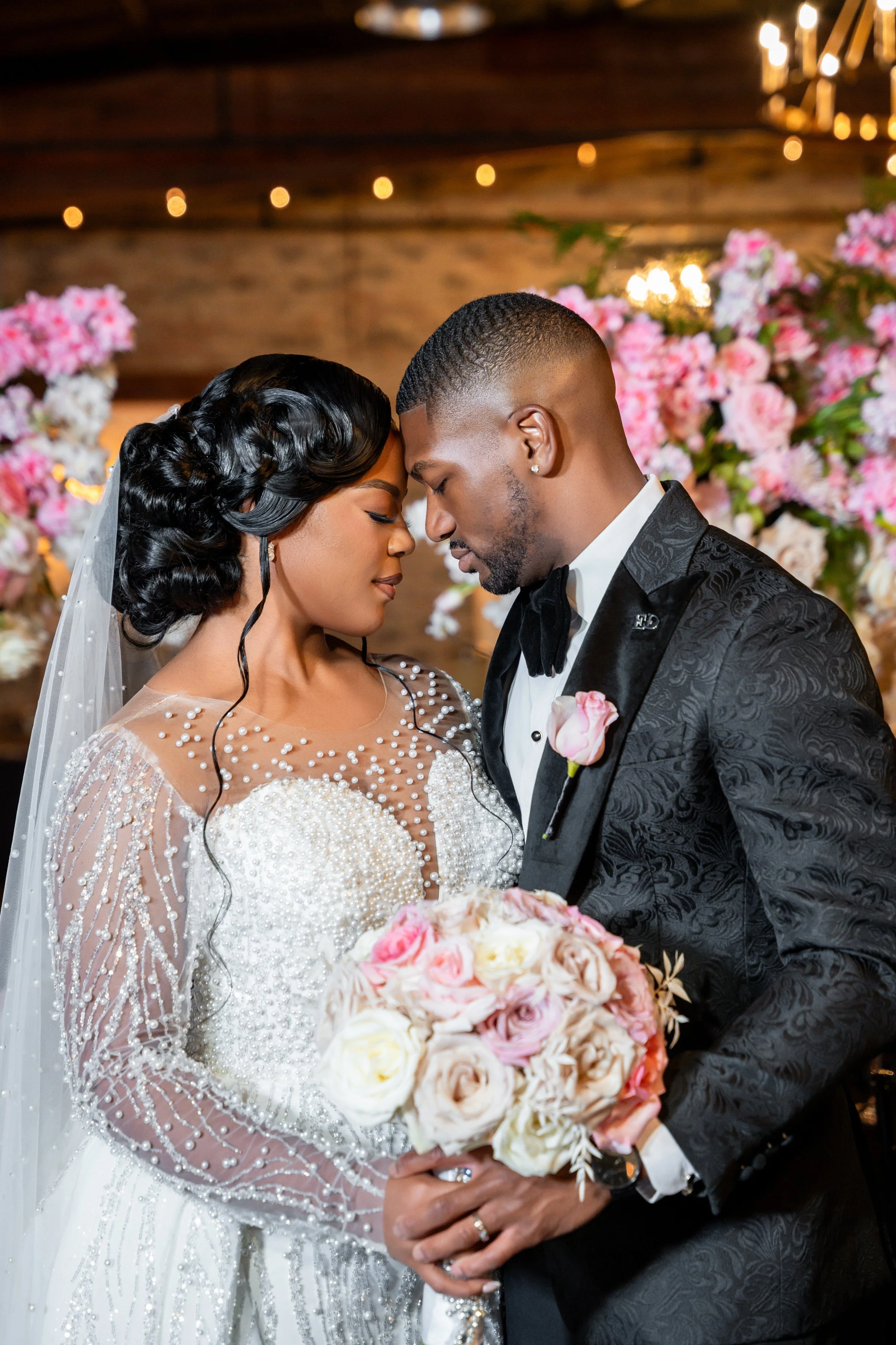 A bride and groom with foreheads touching, dressed in wedding attire, holding a bouquet of pink, white, and cream roses, in a decorated indoor venue.