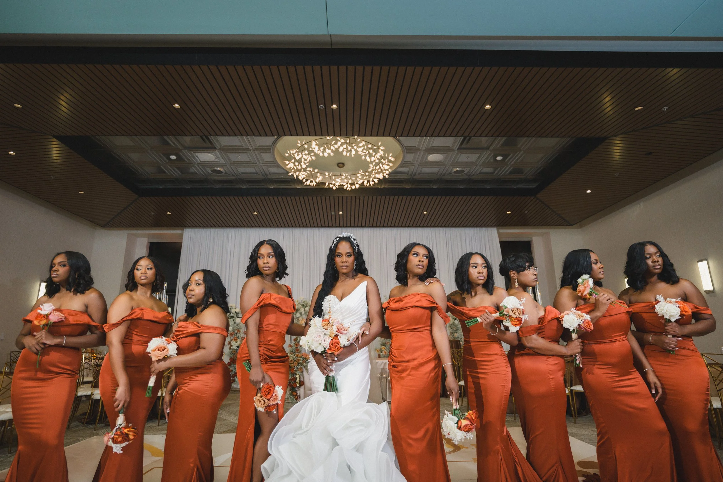 A group of women, some in matching orange dresses and one in a white wedding gown, holding flowers during a wedding ceremony in an indoor venue with a decorative ceiling and a chandelier.