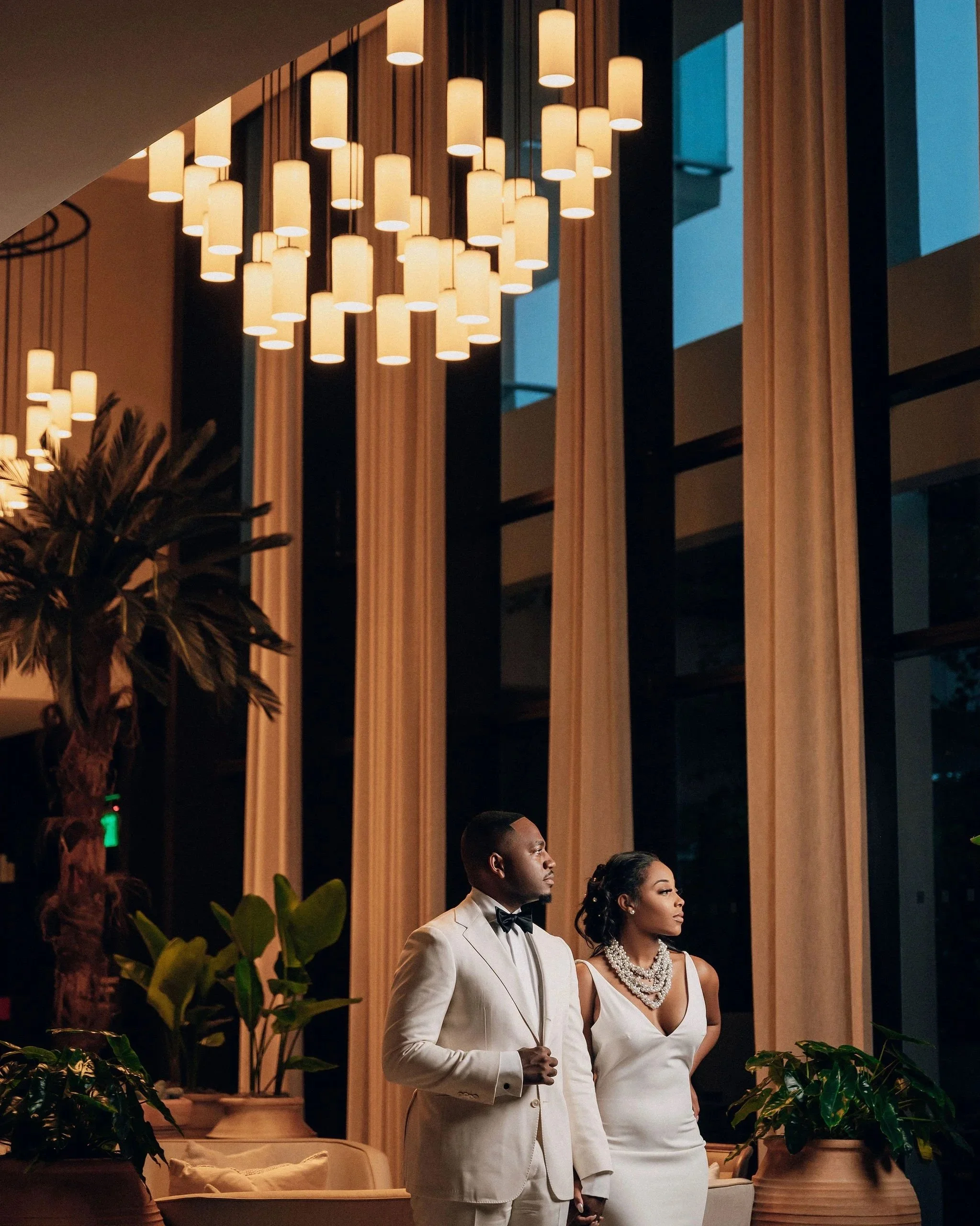 A man in a white tuxedo and a woman in a white dress with pearl jewelry standing indoors at night with warm lighting and decor, including wooden slats, plants, and lounge furniture.