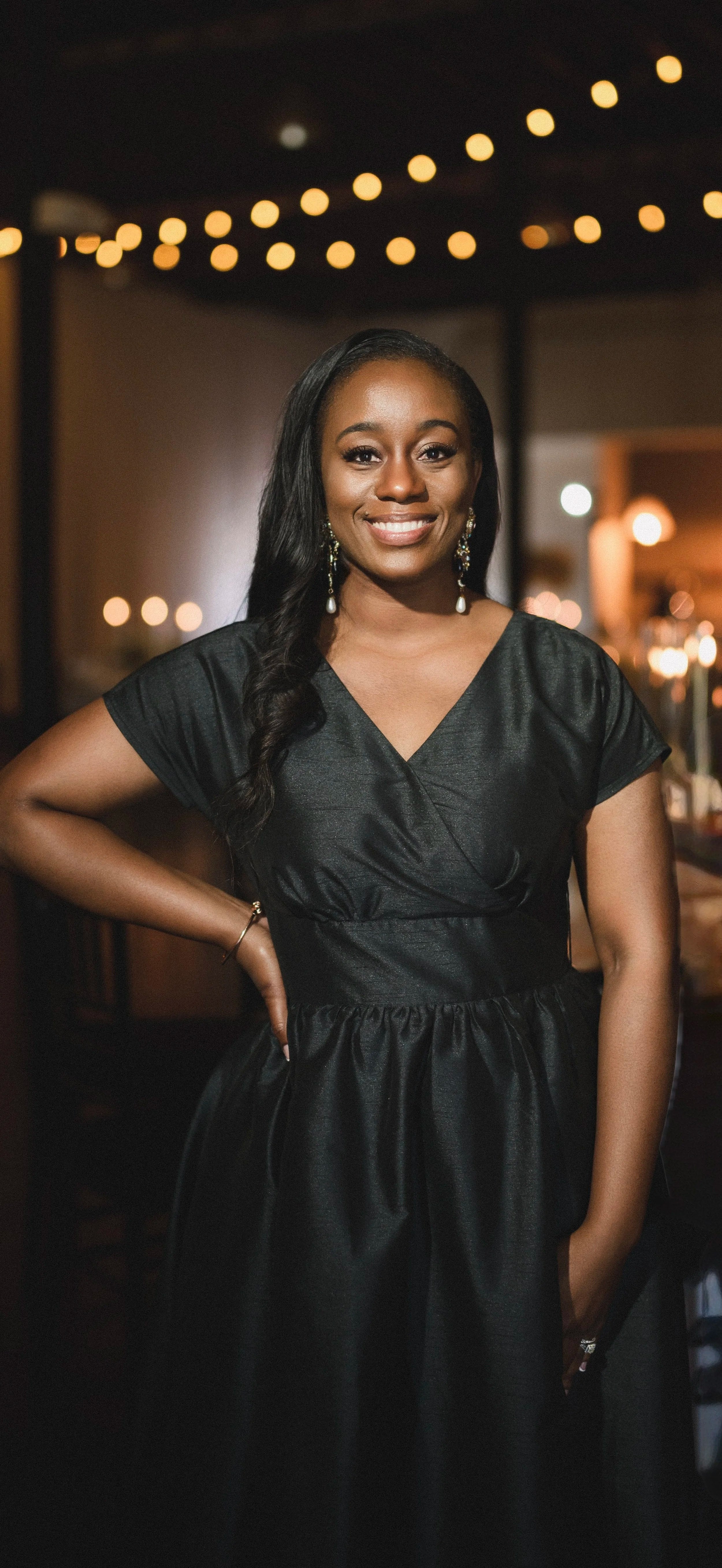 A woman smiling and posing in a black satin dress with earrings, standing in a dimly lit indoor setting with string lights overhead.