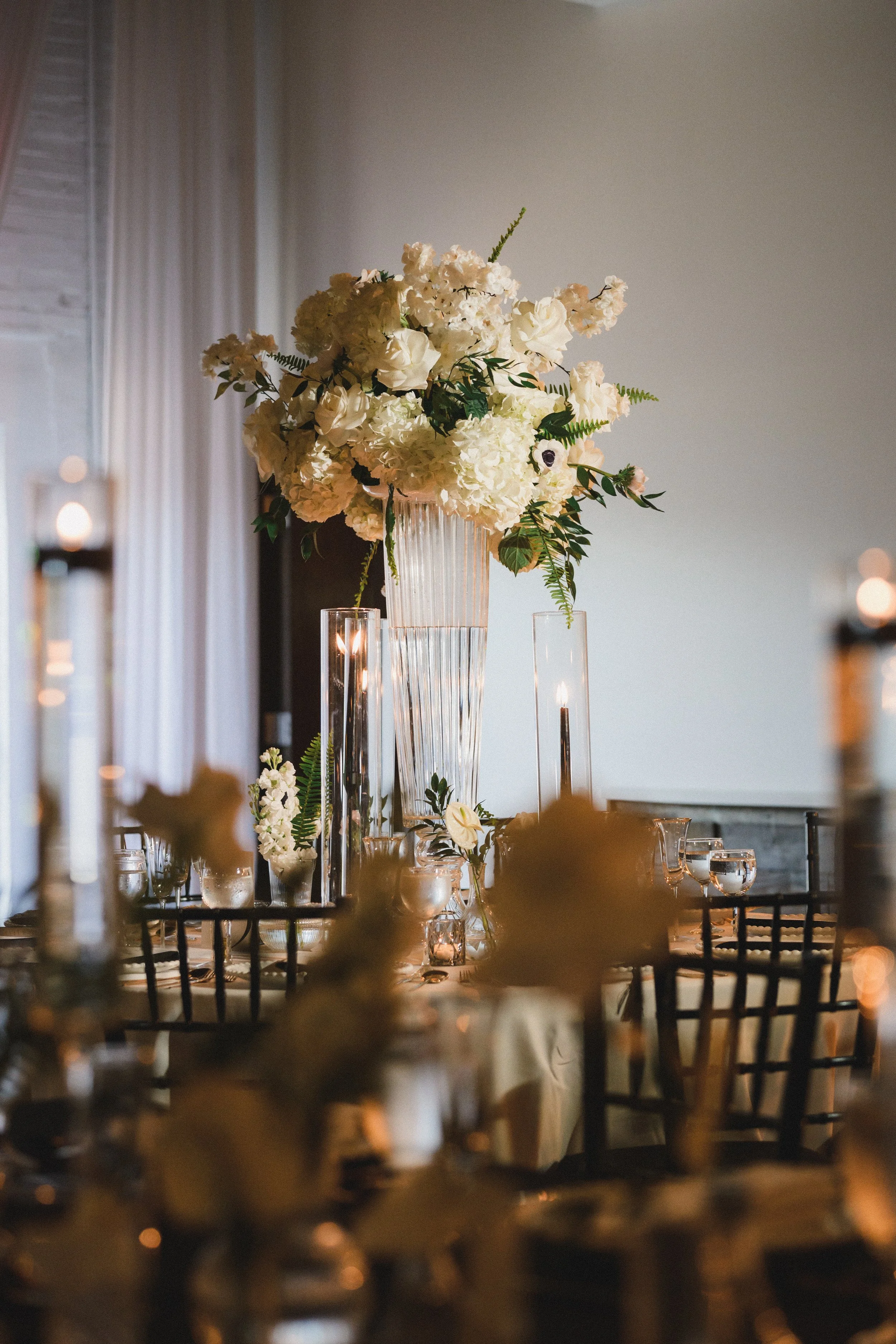 Elegant table setting with a large floral centerpiece featuring white flowers and greenery, surrounded by candles in glass holders and black chairs, likely for a wedding or formal event.