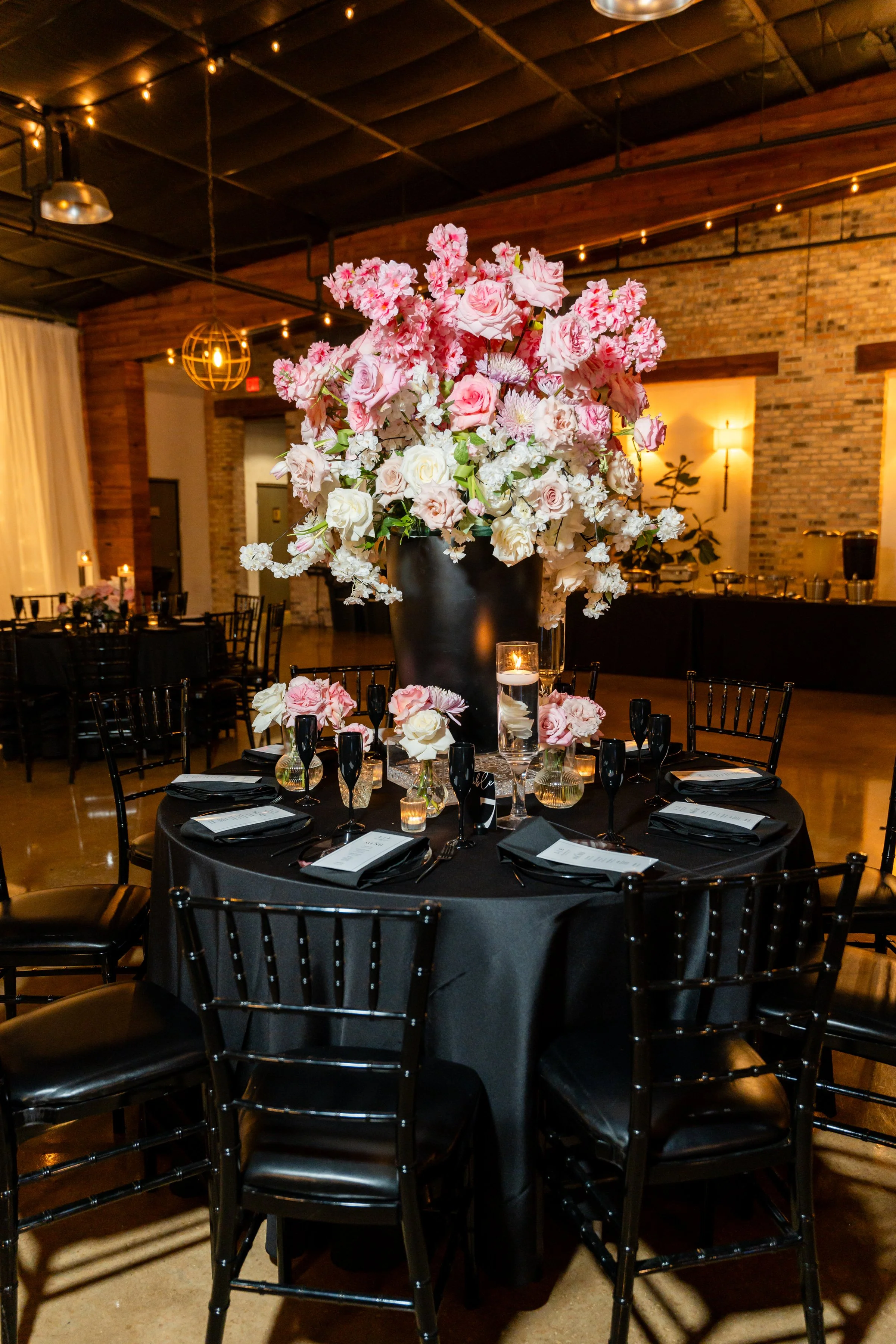 A decorated round table with a black tablecloth, set for a formal event with black plates, black napkins, and glassware. The centerpiece is a large black vase filled with pink, white, and blush flowers, including roses and other blooms, with small ca
