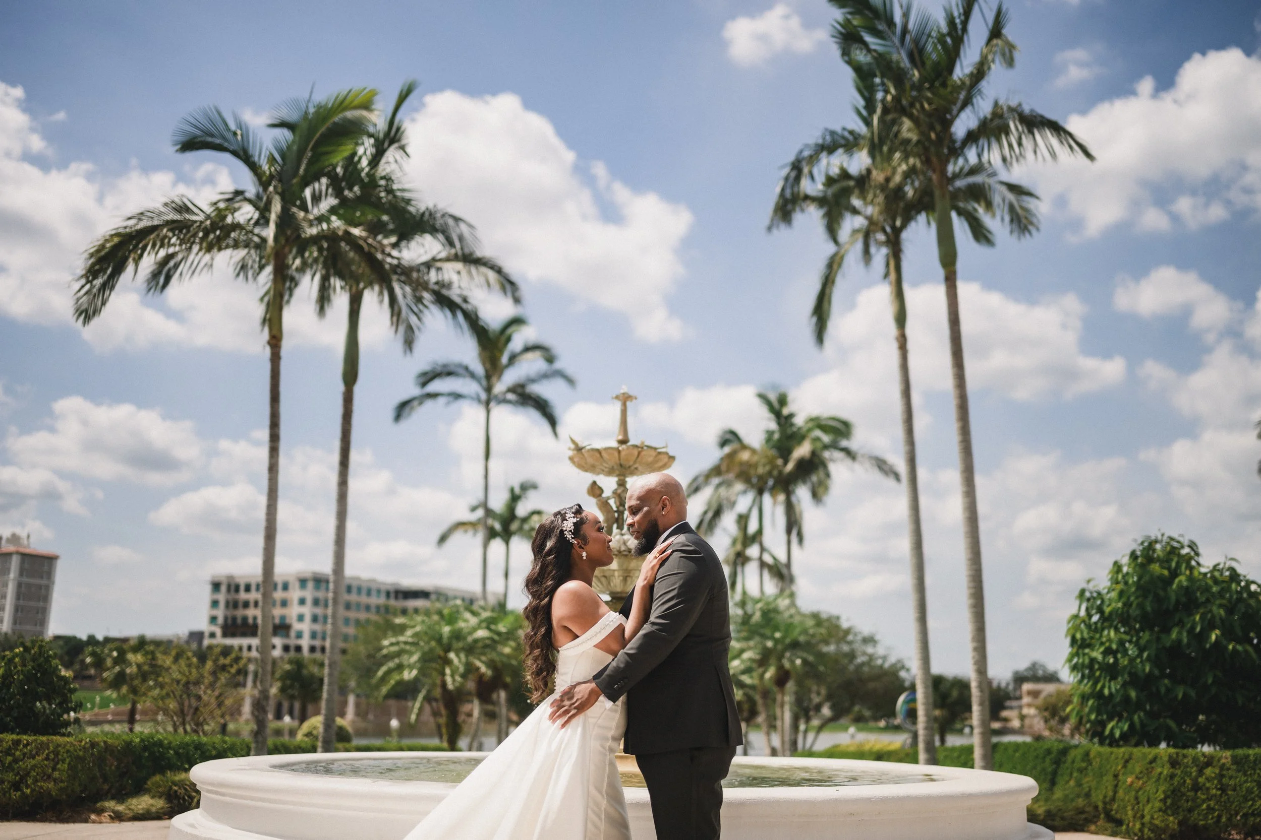 A bride and groom stand close together outdoors, touching foreheads, with palm trees and a blue sky in the background.