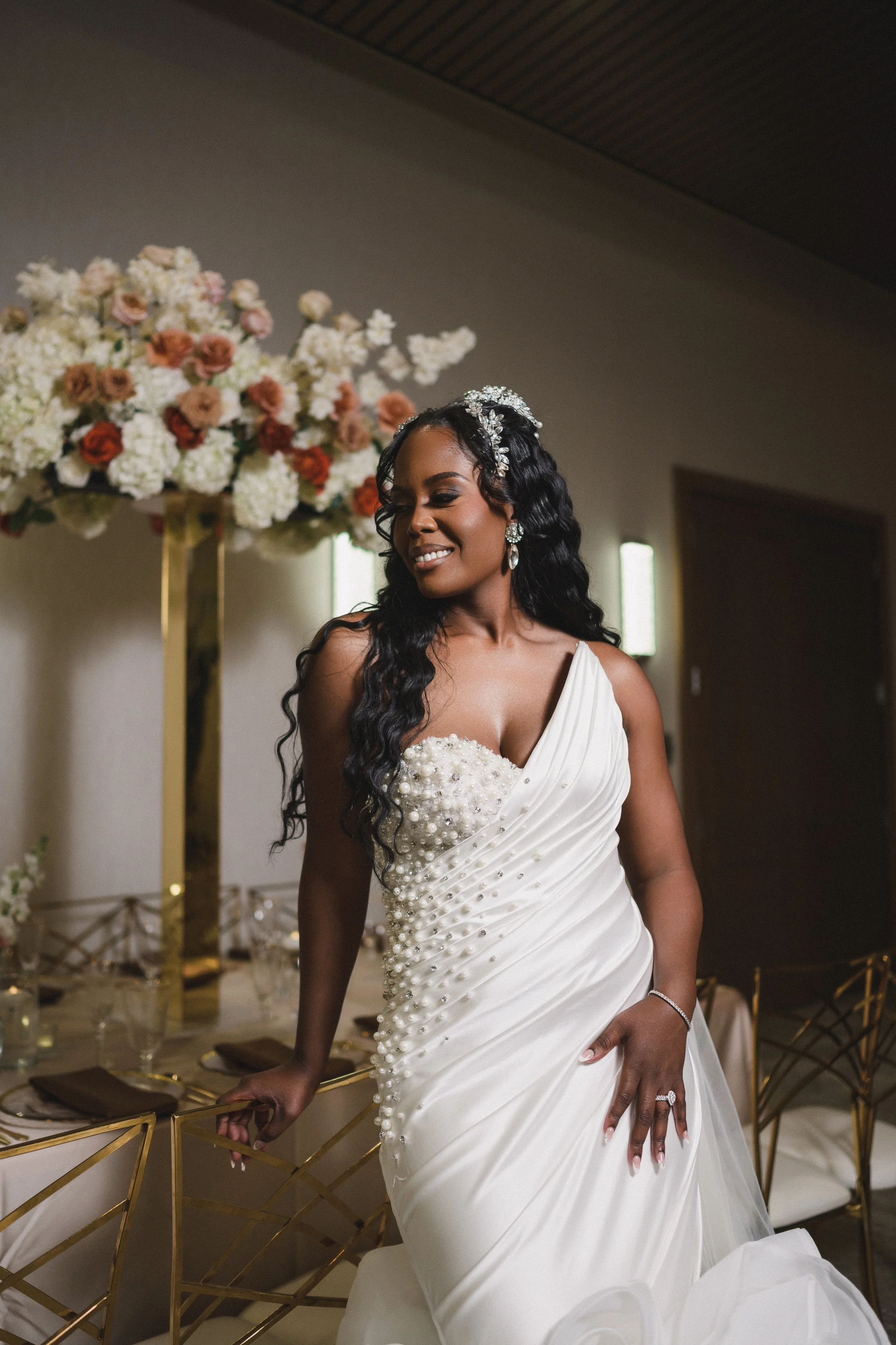 Bride with long black wavy hair wearing a white gown with pearl and jeweled embellishments, standing in a decorated room with floral arrangements.