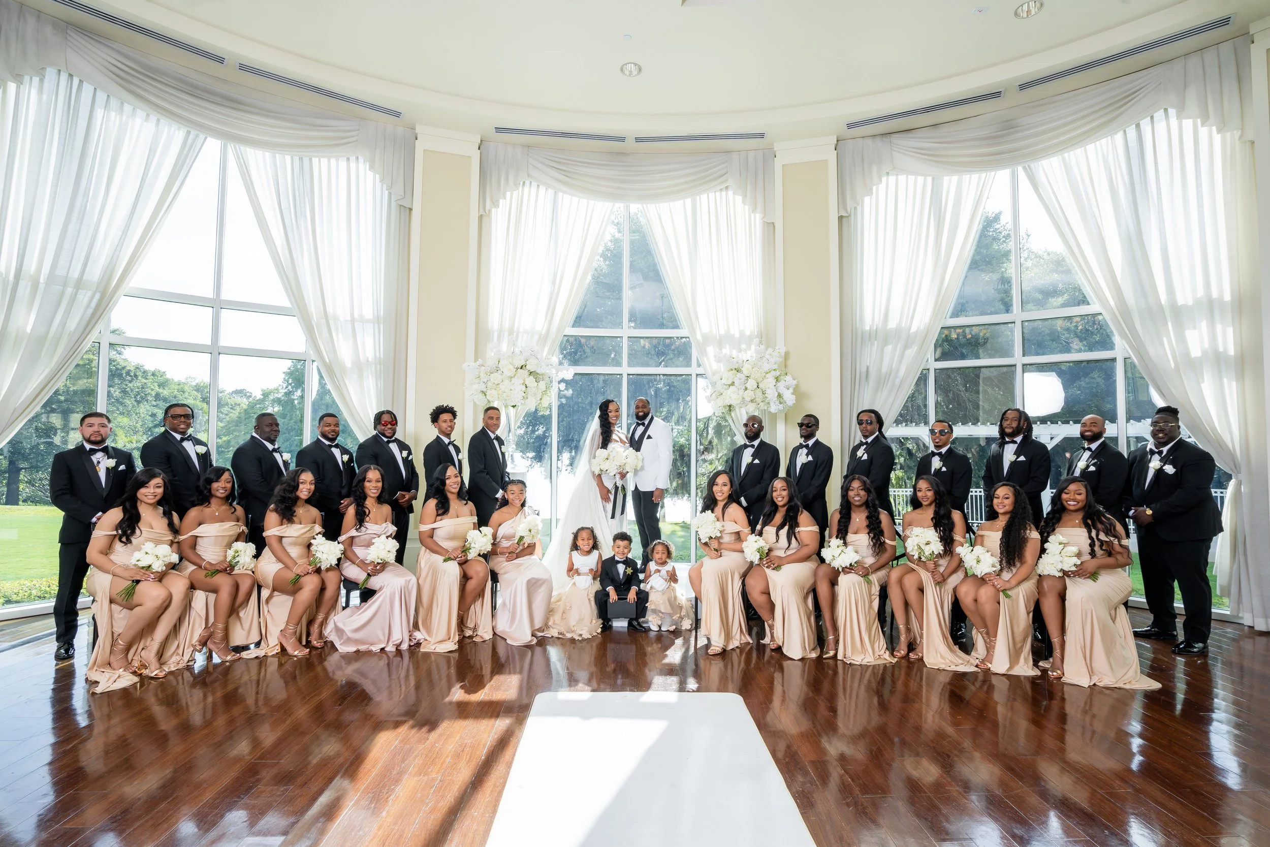 A large wedding party posing indoors with a bride and groom in the center, surrounded by bridesmaids and groomsmen, in a decorated wedding venue with large windows and white curtains.