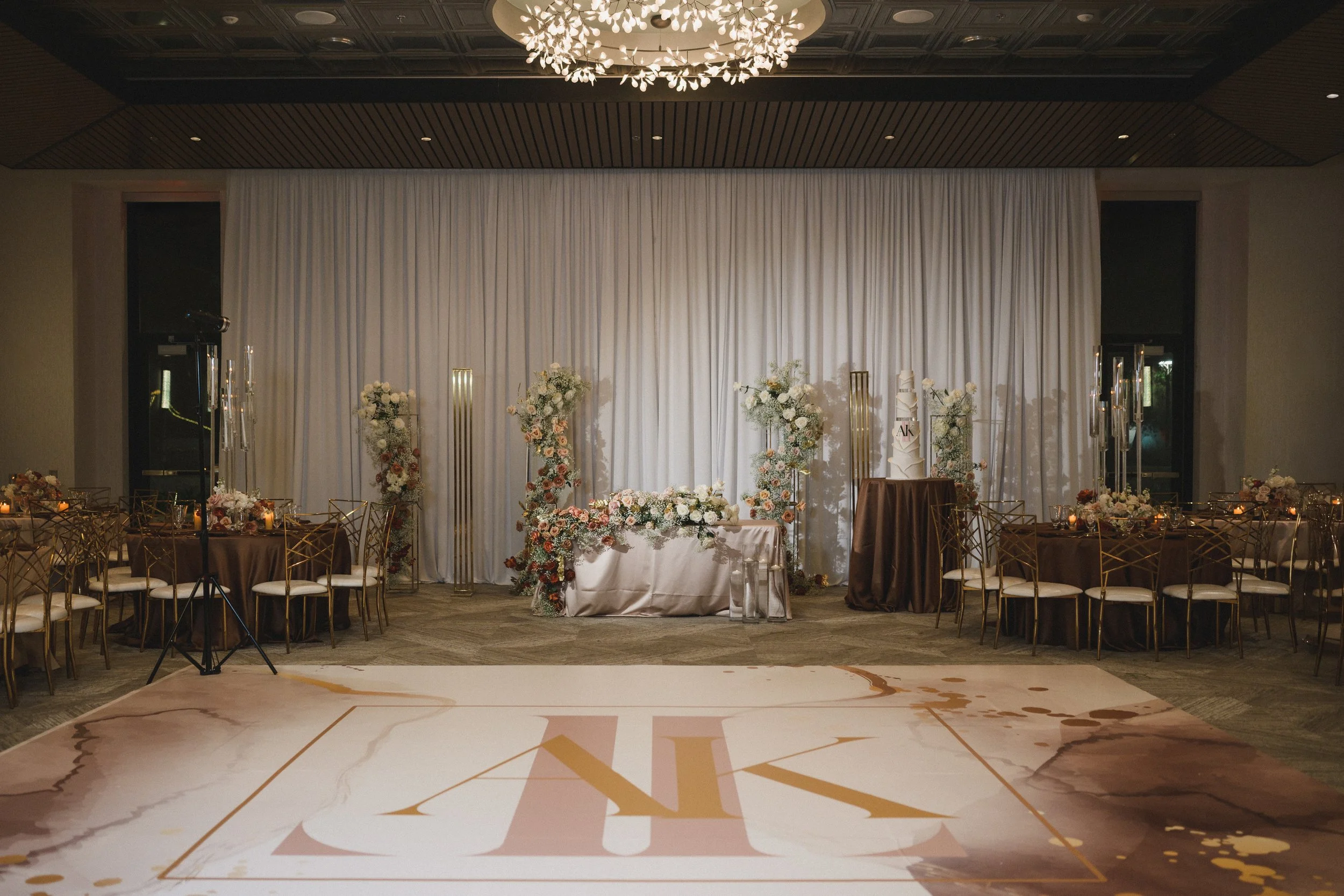 Wedding reception setup with floral arrangements and a tiered wedding cake on a table in front of a white curtain backdrop.
