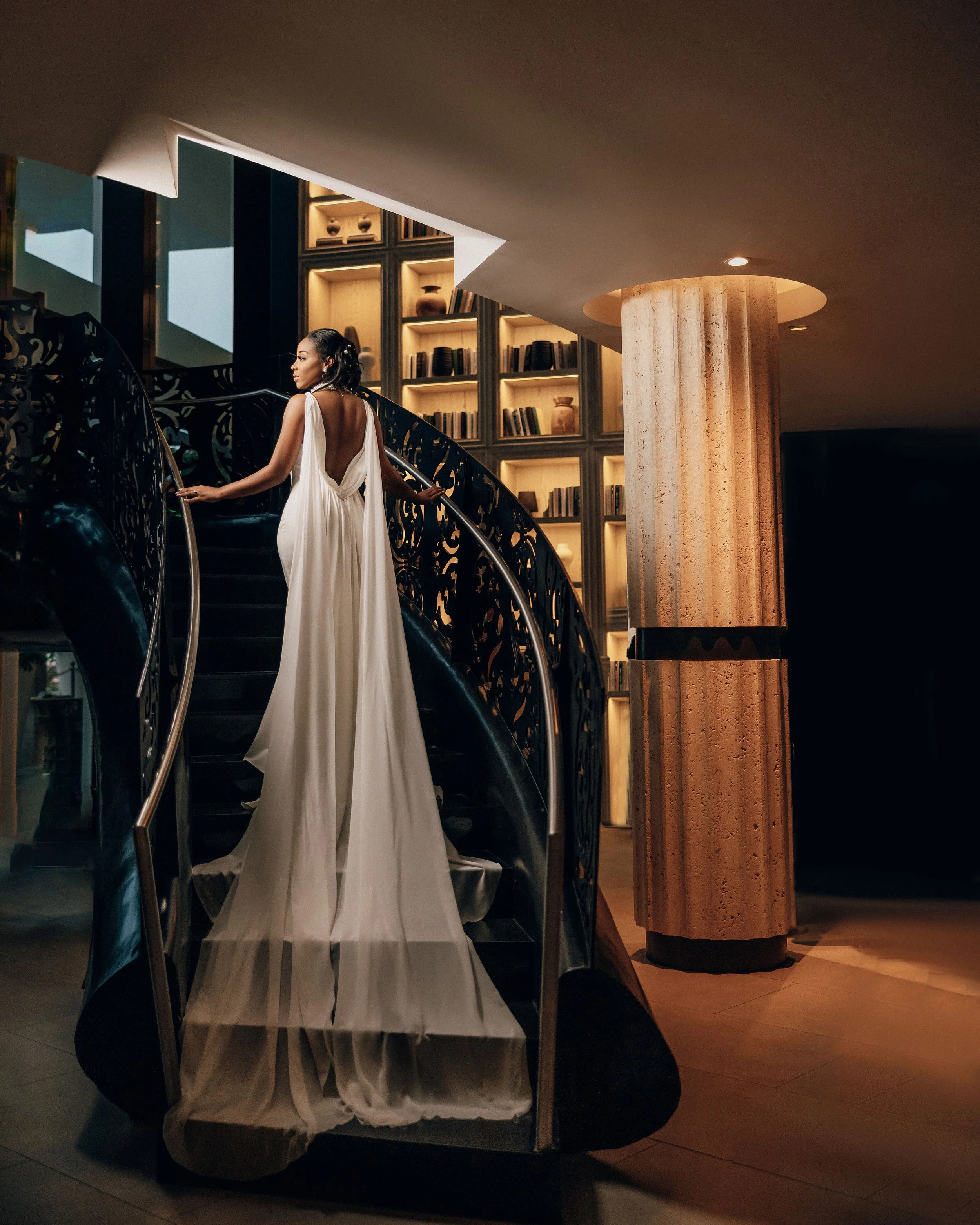 A woman in a white backless gown walking up a spiral staircase with intricate black metal railing in a dimly lit modern interior space, featuring a marble column and built-in bookshelf with various decorative objects.