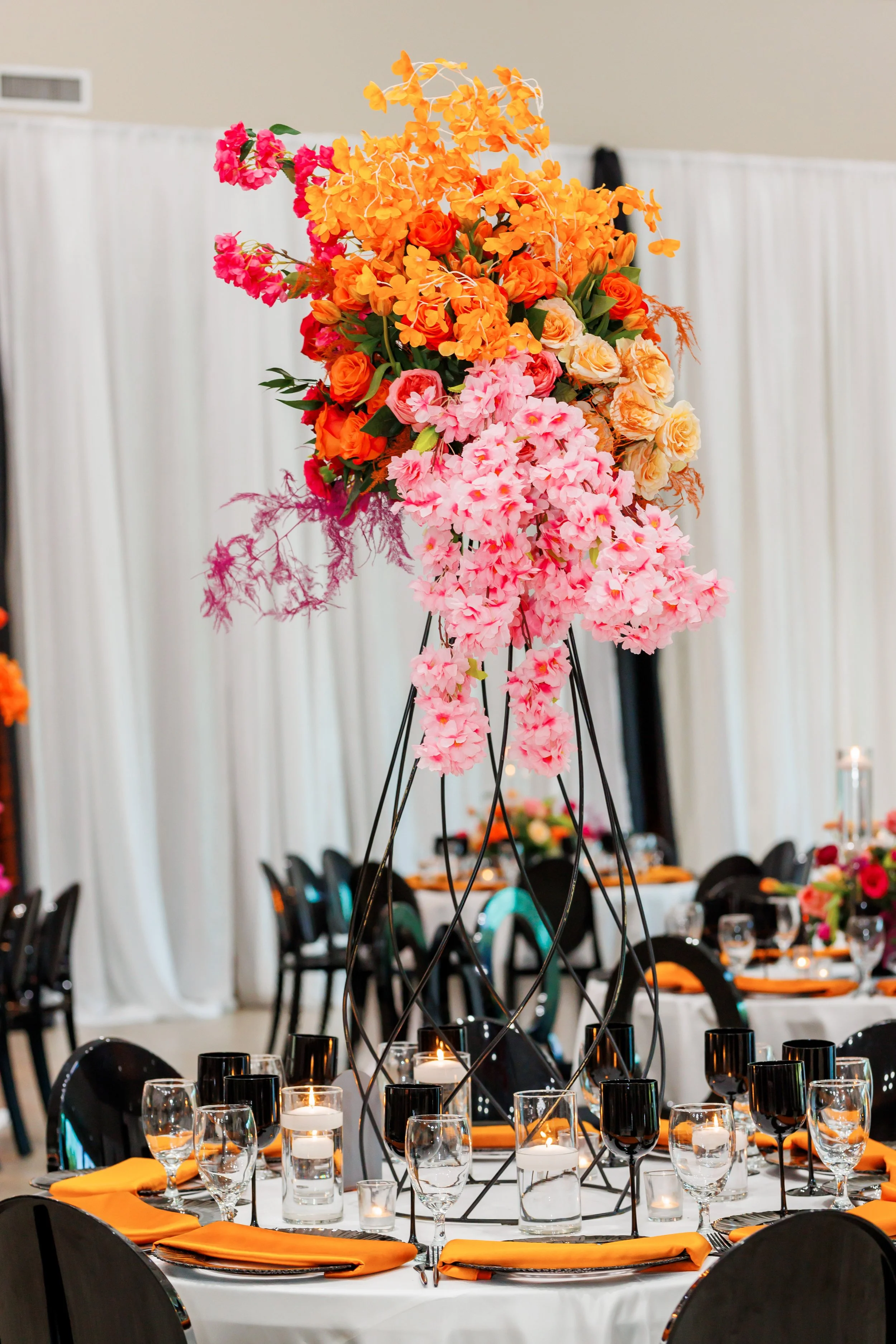 A tall floral centerpiece with orange, pink, and peach flowers on a table at a formal event with black chairs and candles.