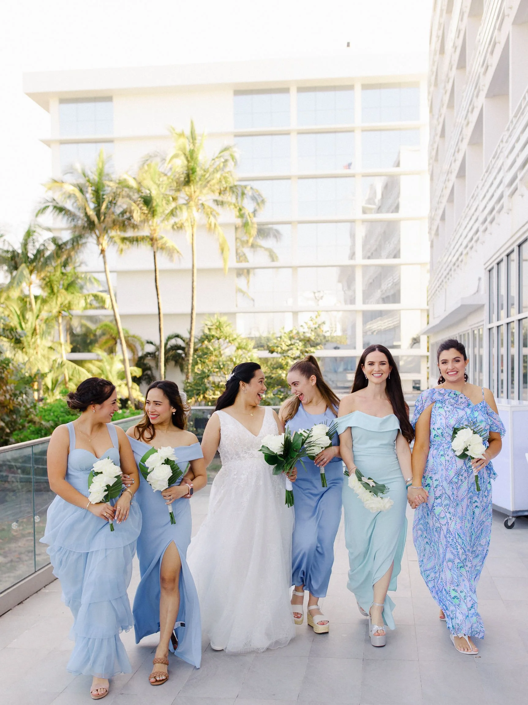 Group of women in pastel dresses, including a bride in a white wedding gown, walking outdoors with city buildings and palm trees in background.