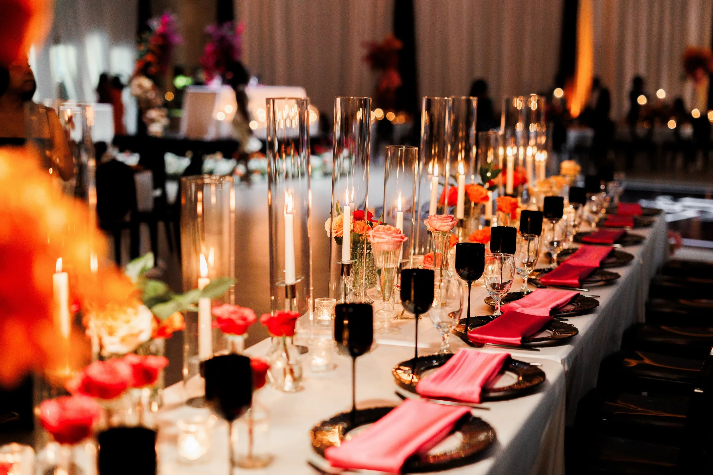 Decorated banquet table set with glasses, plates, pink napkins, candles, and floral arrangements in a dimly lit room for a celebration.