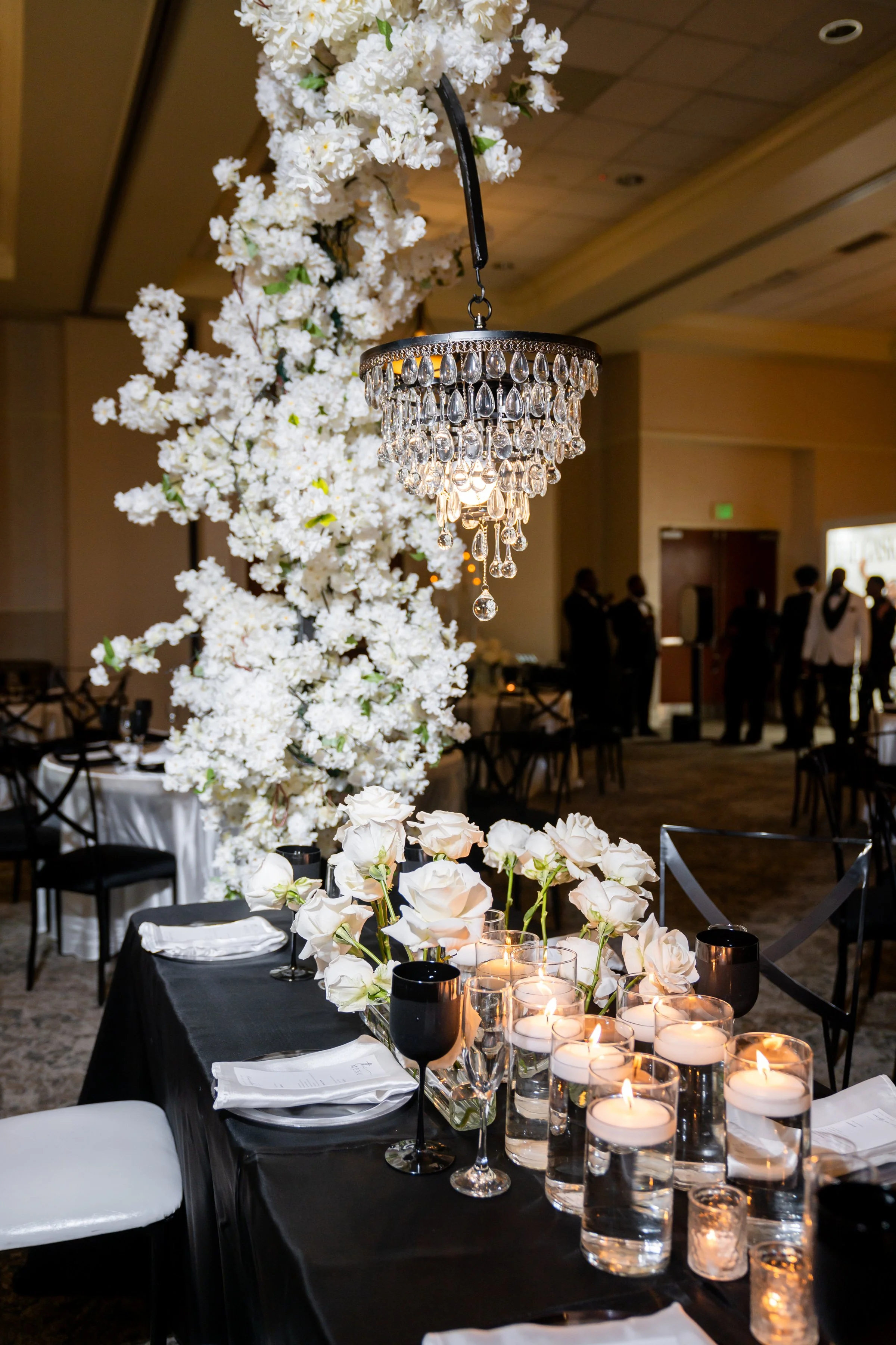 Elegant event table with black tablecloth, white floral centerpiece, candles in glass holders, wine glasses, and black cups, in a decorated event hall with people in the background.