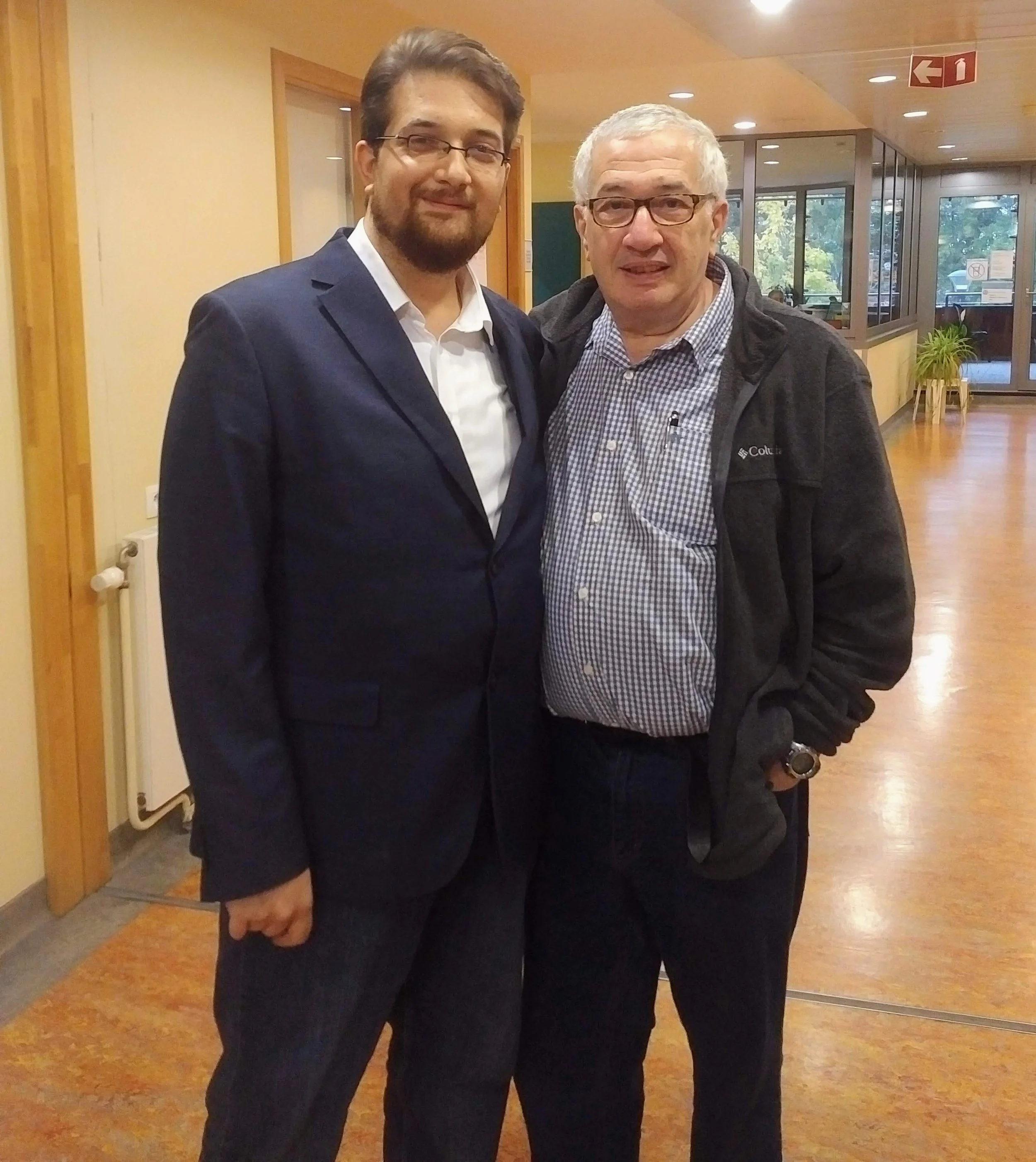 Two men standing close together indoors, one in a suit and the other in a casual jacket, smiling at the camera.