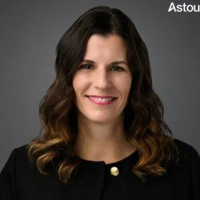 Portrait of a woman with long, wavy brown hair and a friendly smile, wearing a black top with a gold button, against a gray background.