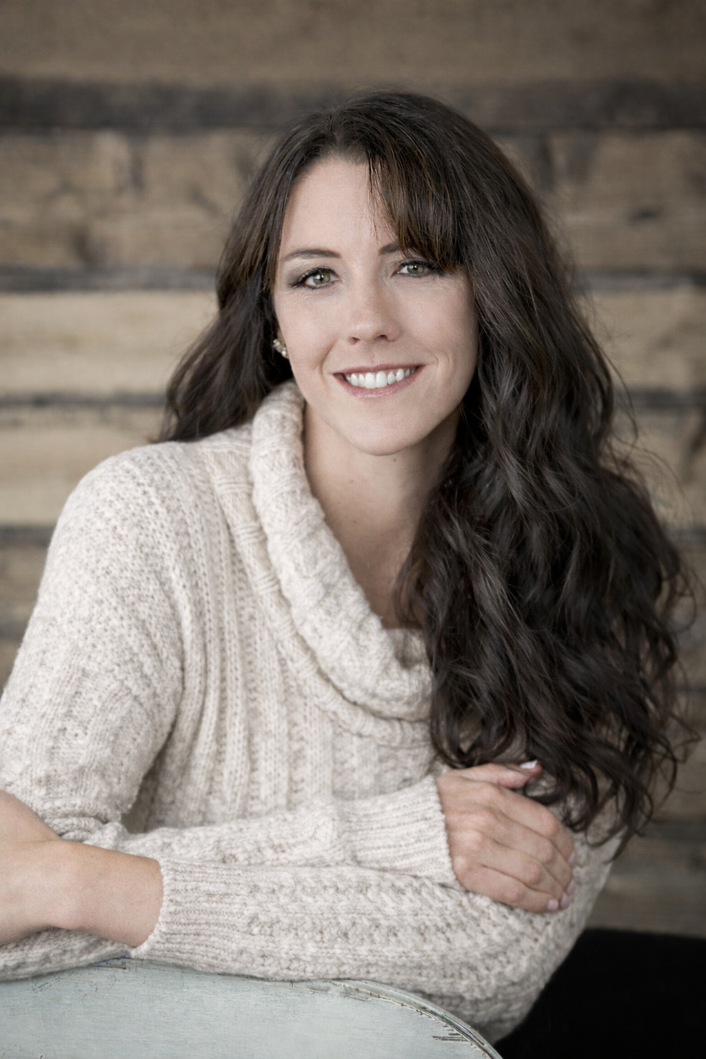 A woman with long dark hair, wearing a black sleeveless top, smiling and sitting with her arms crossed on a table in front of a wooden wall background.