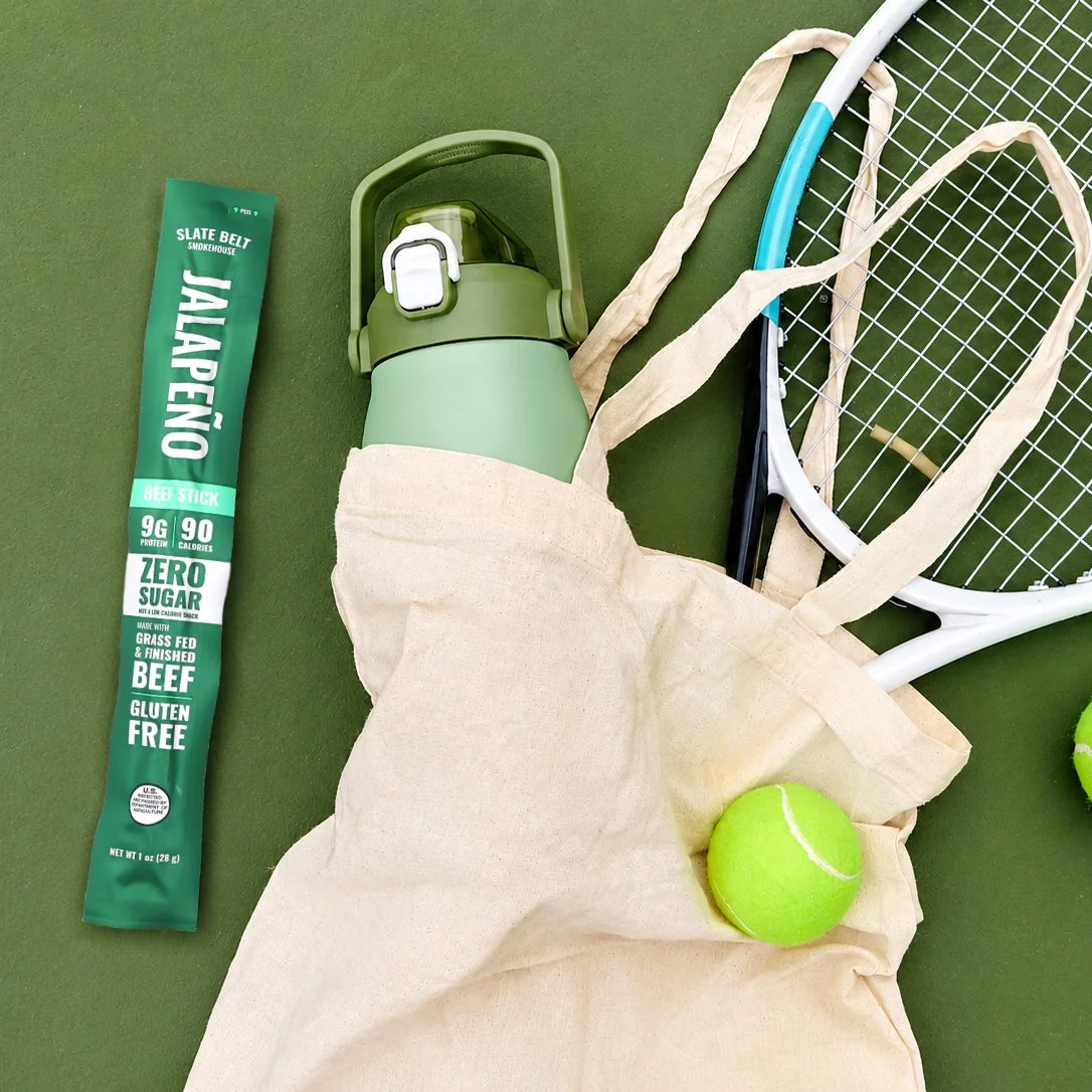 Slate Belt Smokehouse Jalapeño Grass-Fed Zero Sugar Beef Stick with tennis gear, tote bag, and water bottle on a green background.
