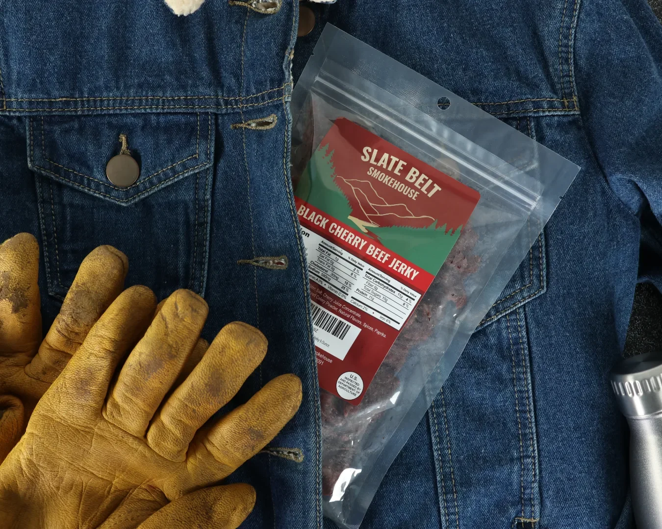 A flat layout of a denim jacket and yellow work gloves with a package of Slate Belt Smokehouse Black Cherry Beef Jerky tucked inside the jacket.