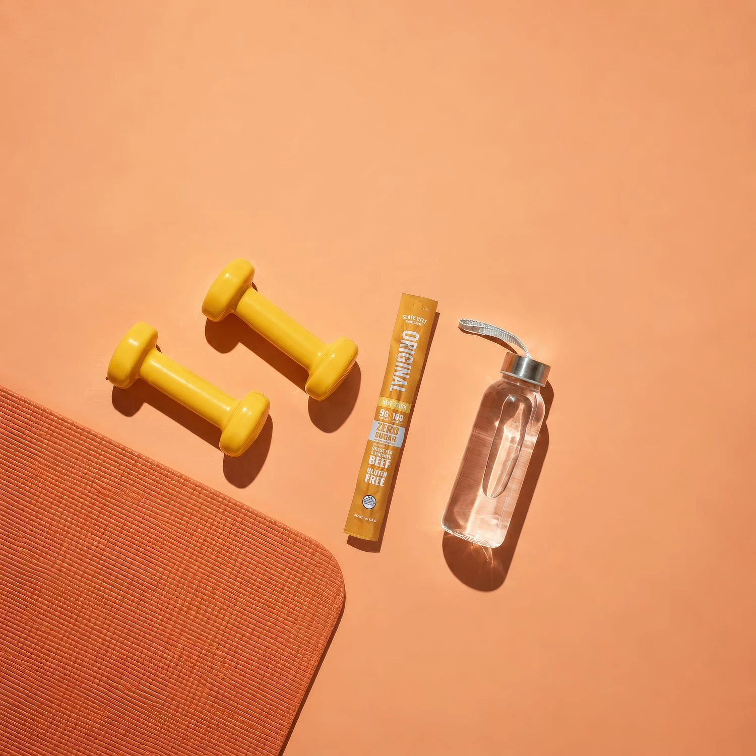Slate Belt Smokehouse Original Grass-Fed Zero Sugar Beef Stick with dumbbells, yoga mat, and water bottle on an orange workout background.