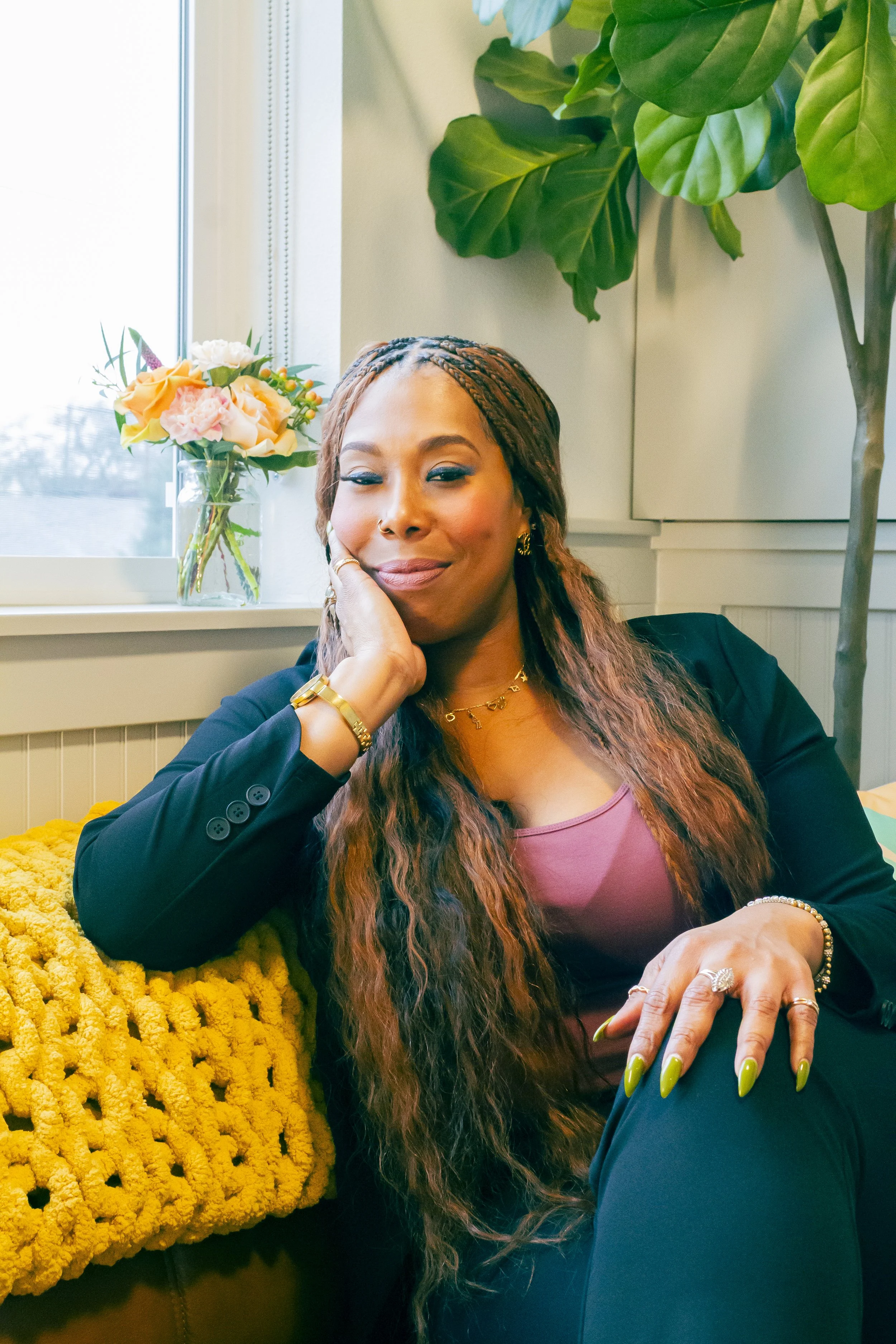 A woman with long black hair and makeup, wearing a black blazer, a gold necklace, and a nose piercing, standing near a body of water with trees in the background.