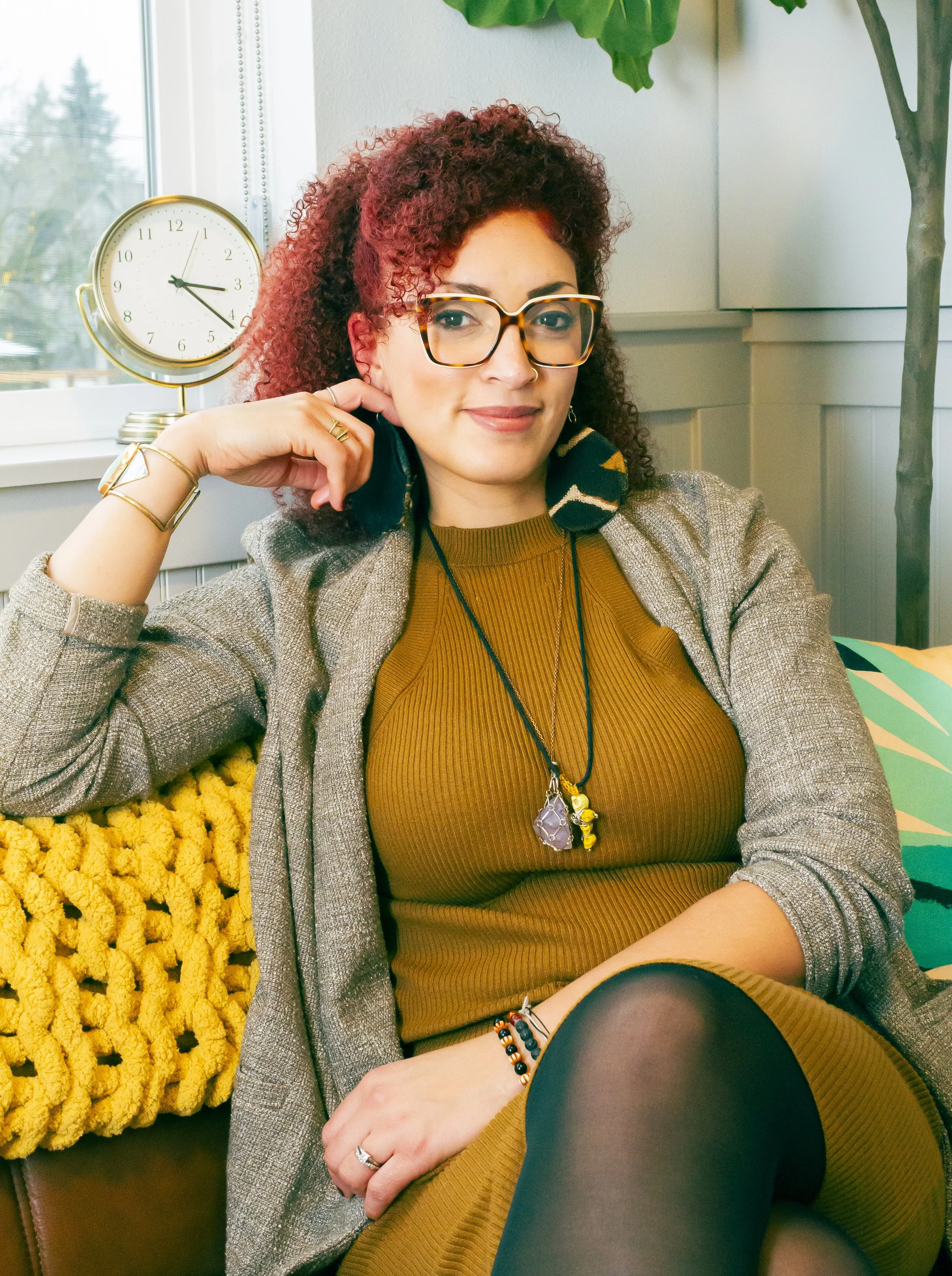 A woman with curly brown hair wearing black glasses, gold earrings, a white shirt, and a mustard-colored cardigan, sitting indoors with green plants in the background.