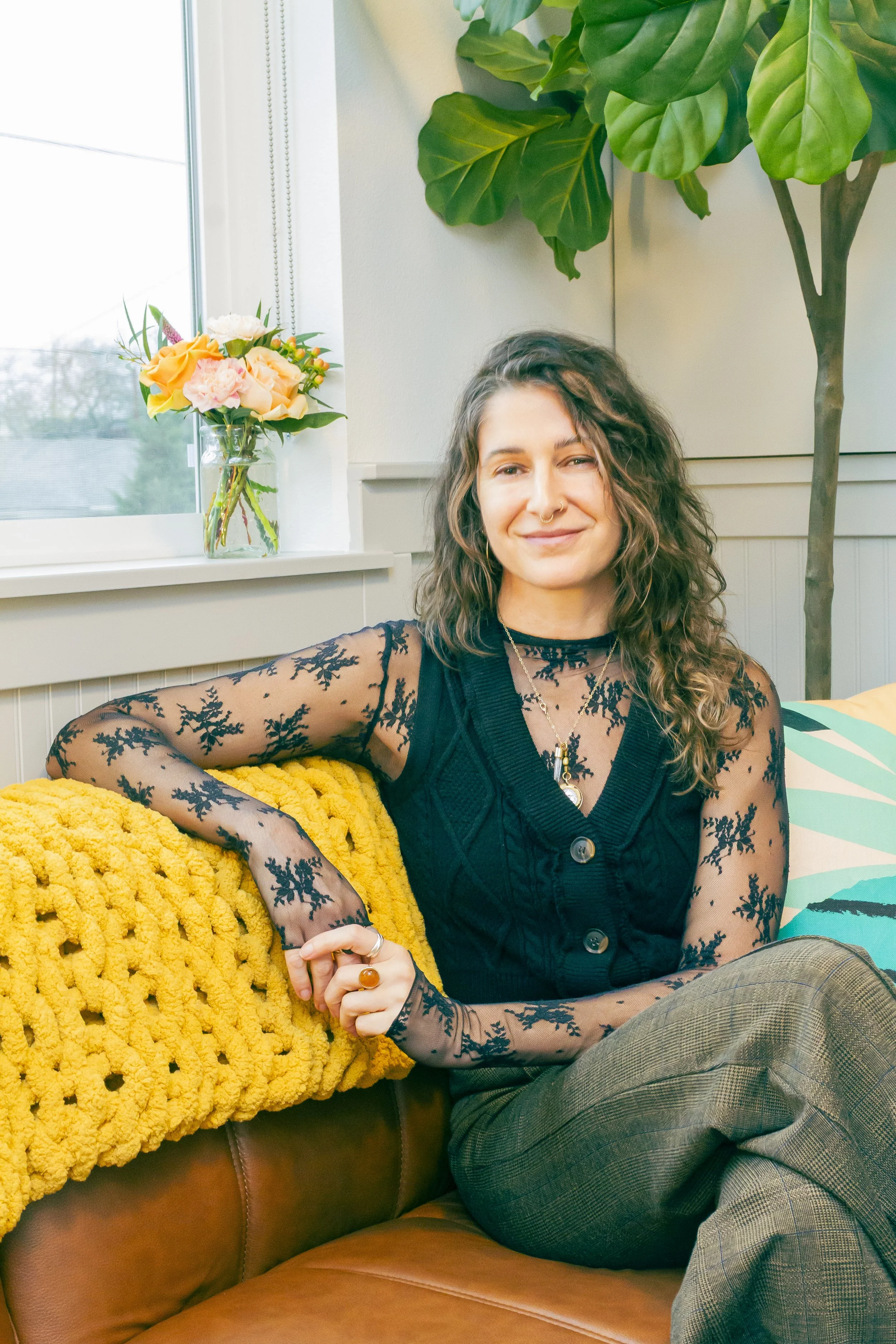 A woman with curly brown hair smiling outdoors during daytime, standing in front of a flowering tree with pink and white blossoms.
