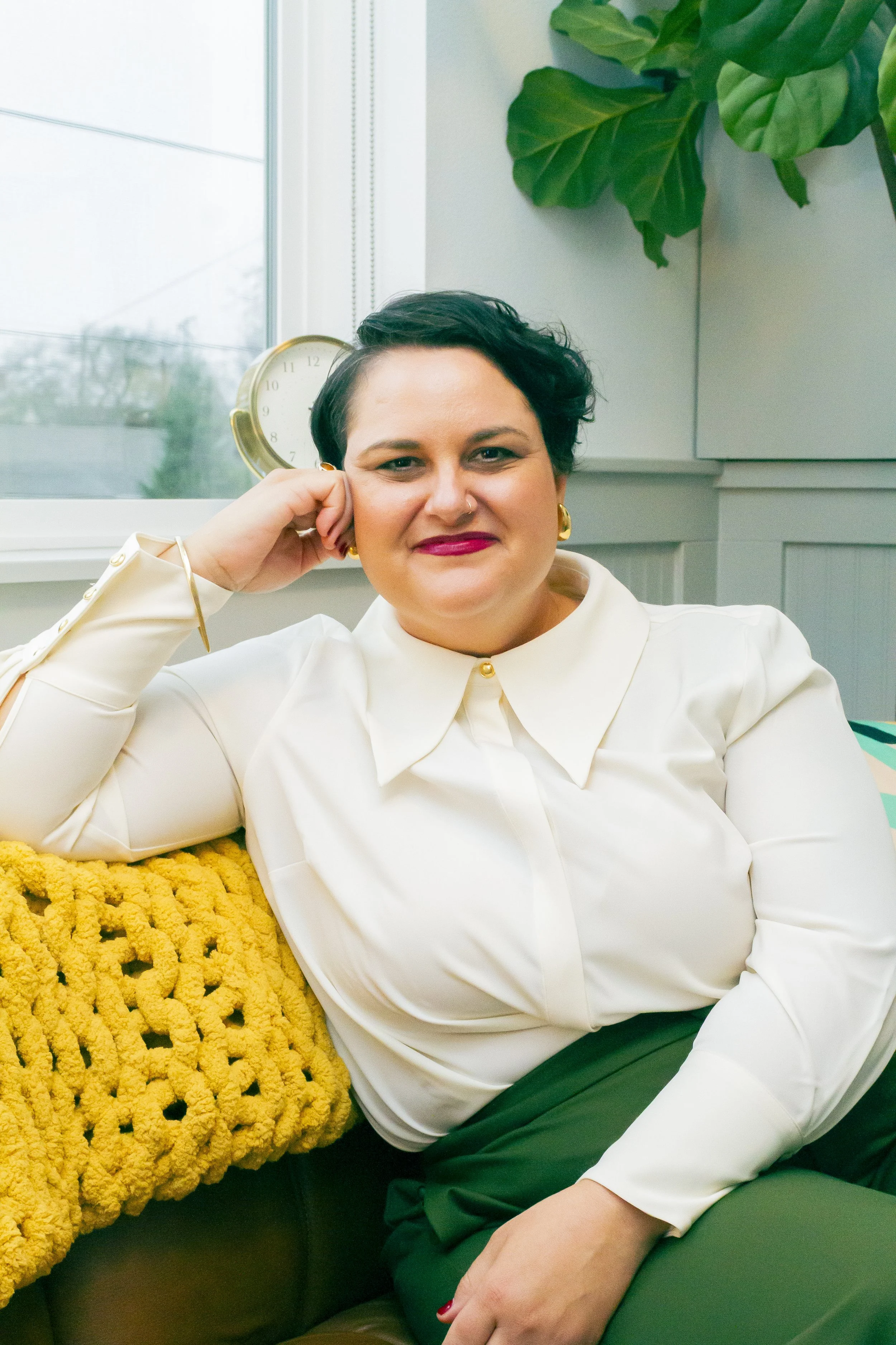 A woman with short dark hair and earrings, wearing a denim jacket and black top, smiling outdoors during sunset.