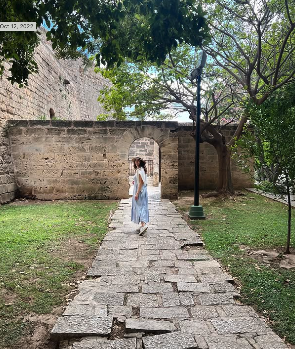 A woman in a long light blue dress and wide-brimmed hat walking on a stone path through an archway in an old brick wall, surrounded by green grass, trees, and a streetlamp.