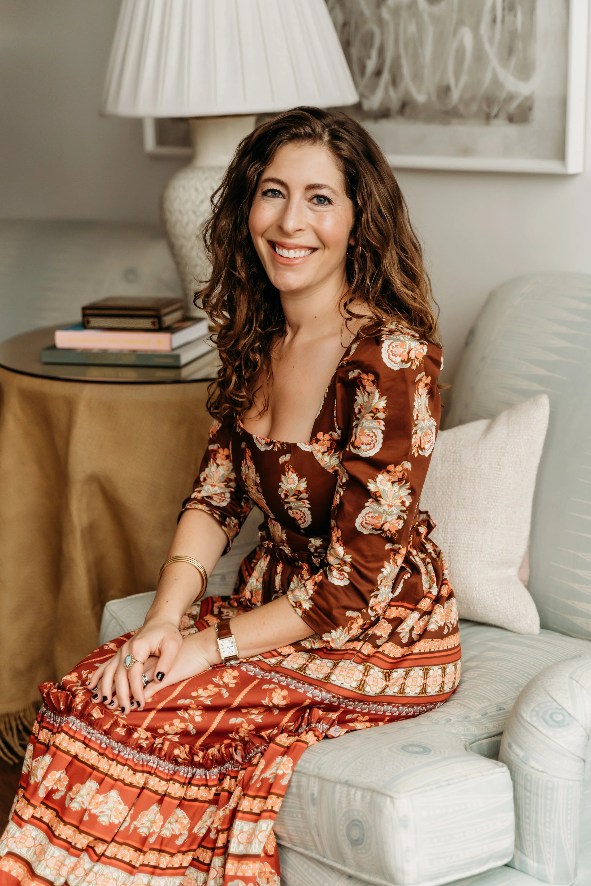 A woman with curly brown hair, smiling and sitting on a chair in a cozy living room, wearing a floral brown dress.