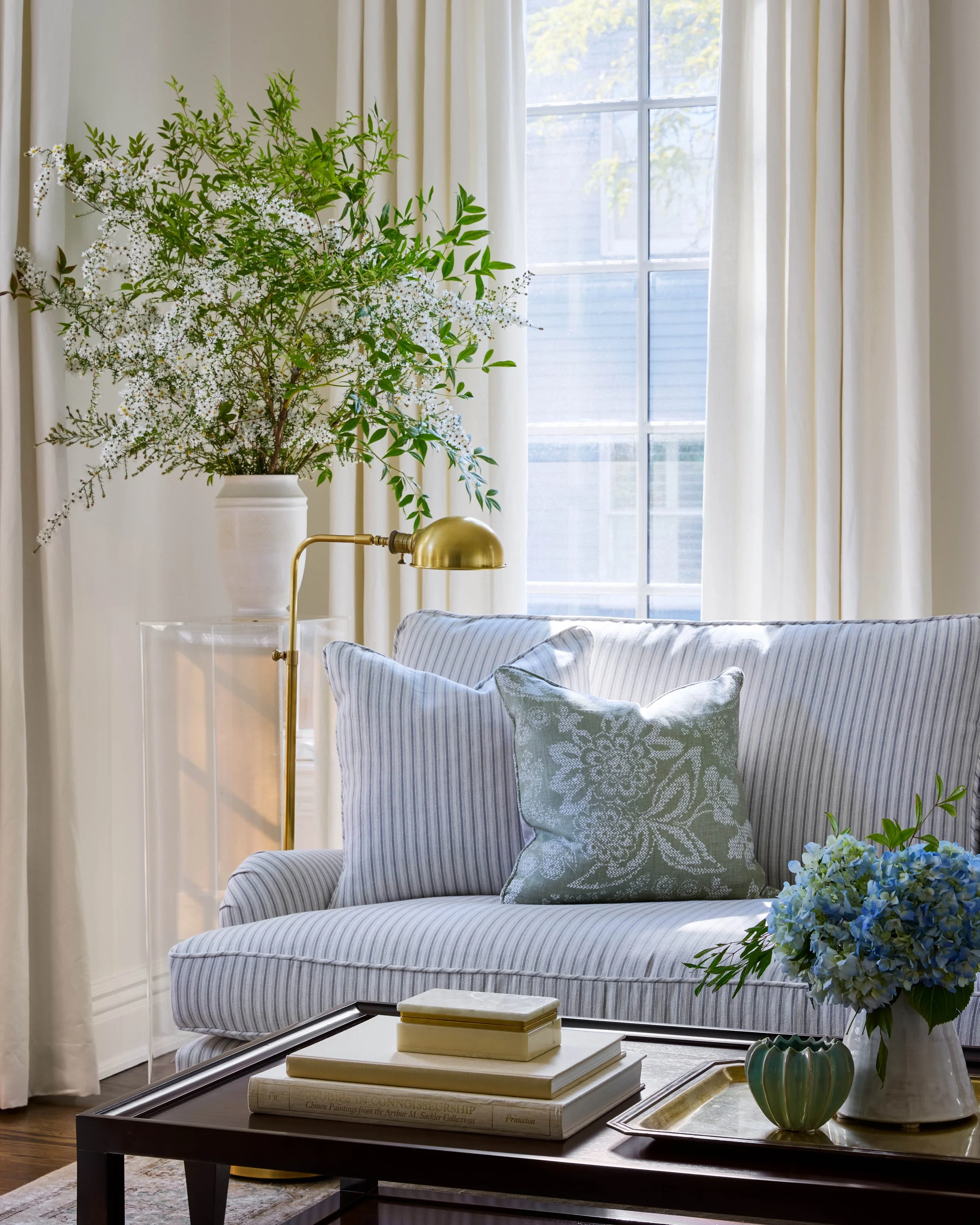 A living room with a light gray and white striped sofa, blue and white throw pillows, a wooden coffee table with books and a green vase with blue hydrangeas, a large potted plant with white flowers near a window with cream curtains, and a brass reading lamp.