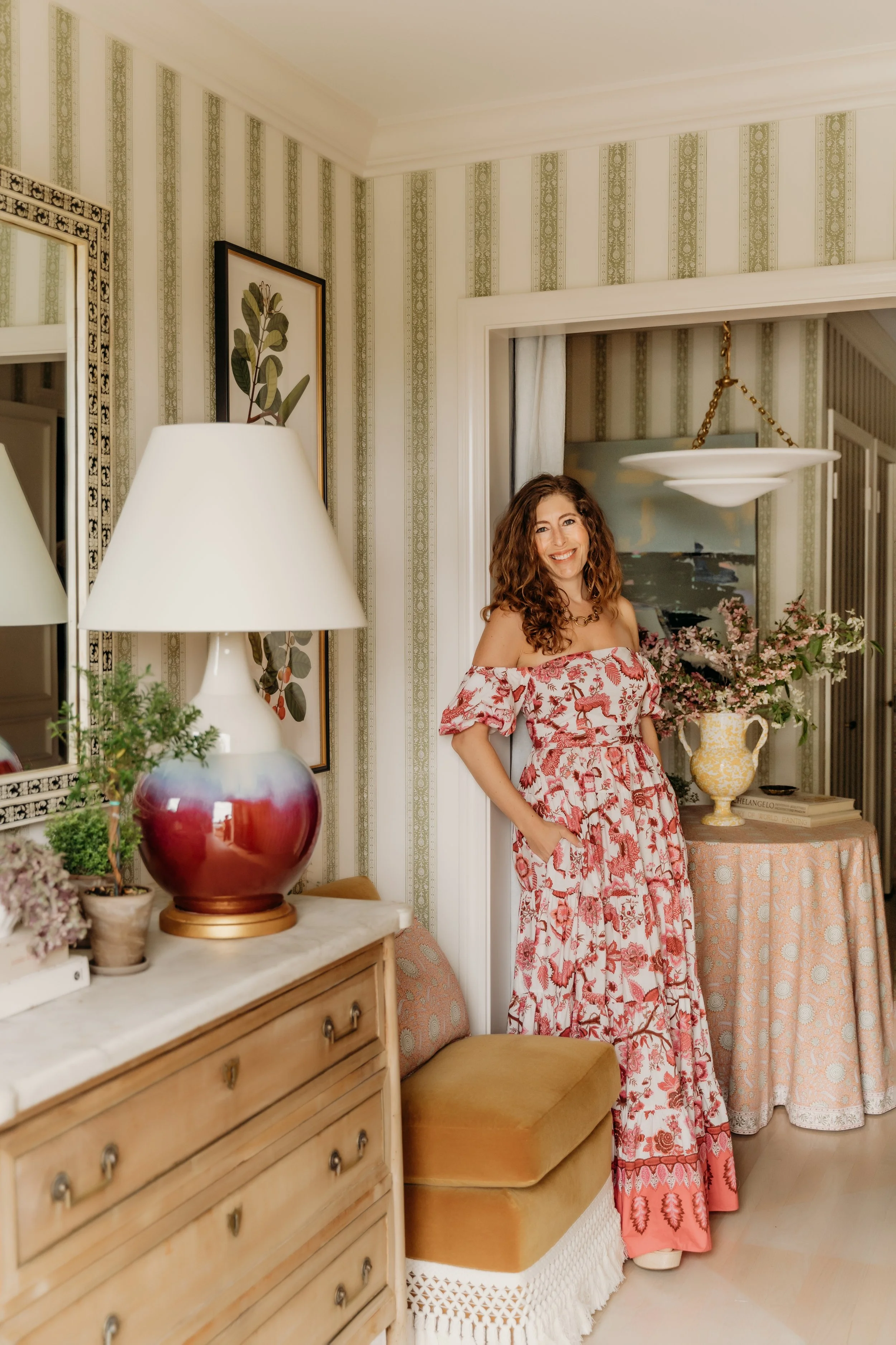 A woman in a floral off-the-shoulder dress standing in a vintage room, smiling, with green and white wallpaper, a table with flowers, and a mirror.