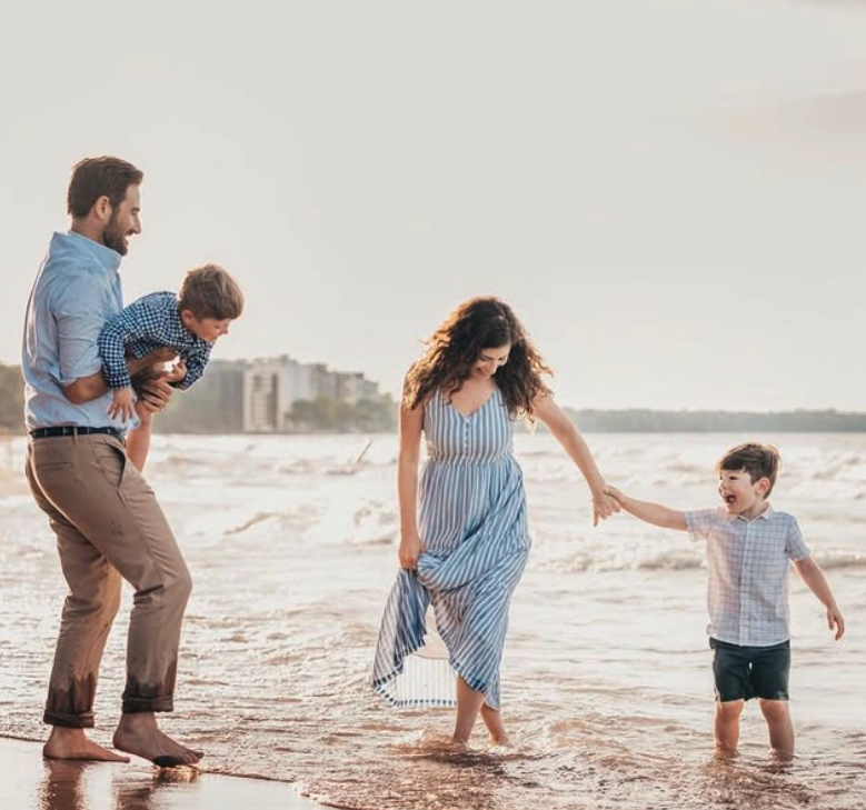 A family of four enjoying time together on the beach, with the father holding a young boy, the mother holding hands with a smiling young boy, all walking in the shallow water.