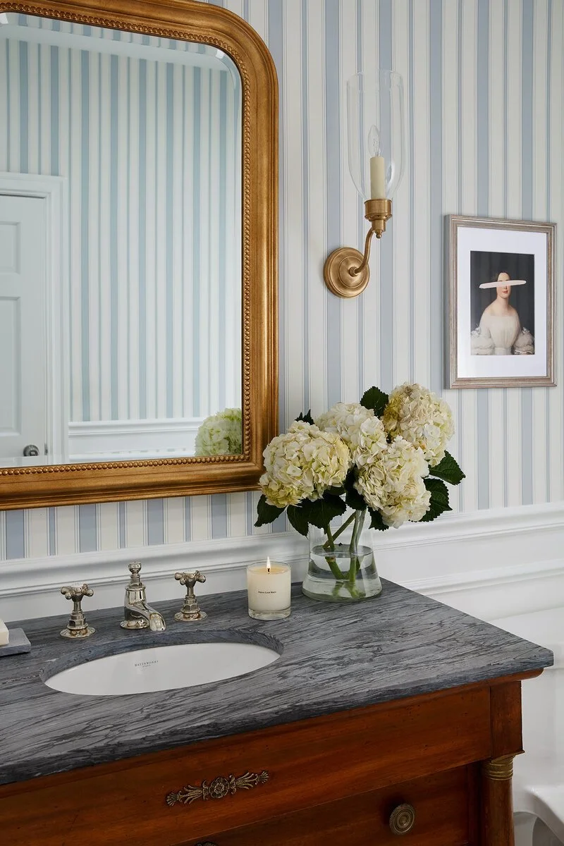 A bathroom vanity with a dark gray marble countertop, a white undermount sink, silver faucets, a glass vase with white hydrangeas, a lit candle, a gold wall sconce with a candle-shaped bulb, a framed portrait of a woman wearing a white dress with a wide-brimmed hat, and a large gold-framed mirror reflected in the wall.