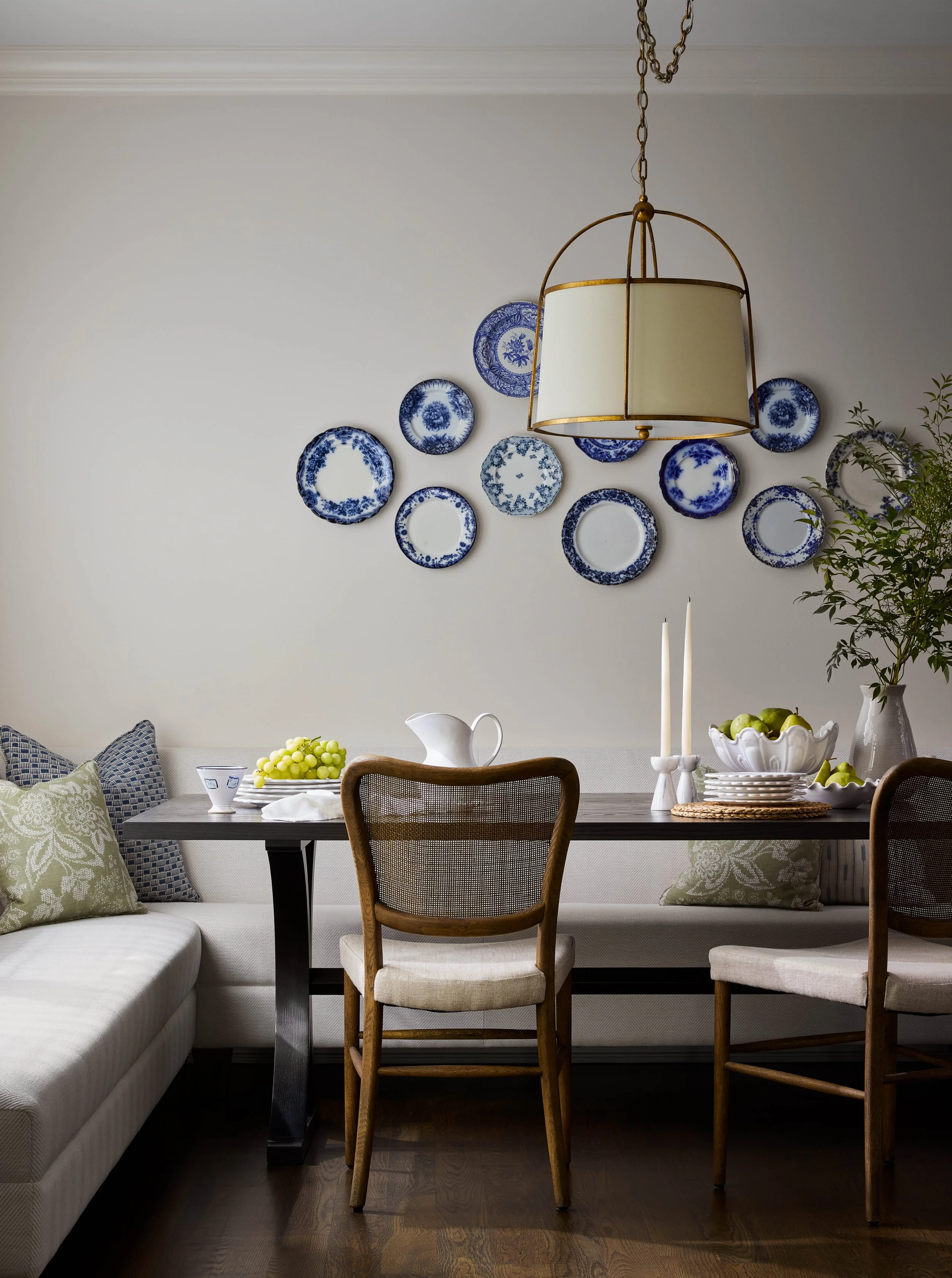 A dining room with a dark wood table, beige upholstered chairs, and a white banquette. The table has a white pitcher, a bowl of green apples, candles, and a small plate of grapes. Blue and white decorative plates are mounted on the wall, with a brass chandelier hanging above.