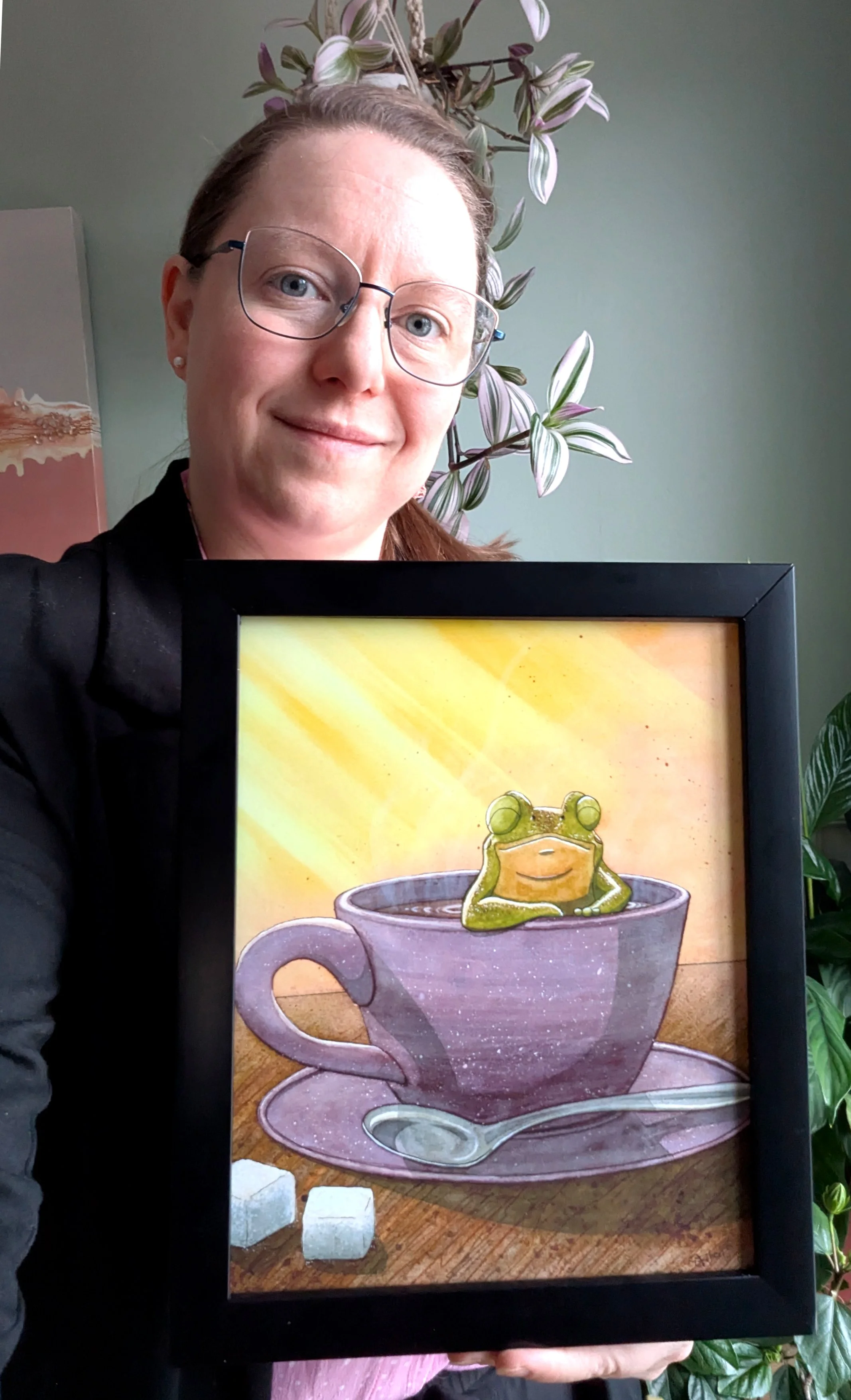 Une femme souriante portant des lunettes tient un cadre avec une peinture d'une grenouille assise dans une tasse de café, dans un décor intérieur avec des plantes en arrière-plan.