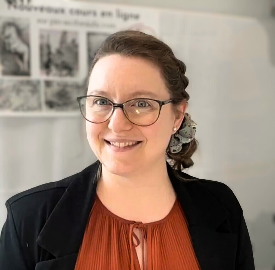 Jeune femme souriante portant des lunettes, cheveux attachés avec un foulard, tenue professionnelle, en intérieur avec un mur blanc et une affiche ou un panneau en gris en arrière-plan.