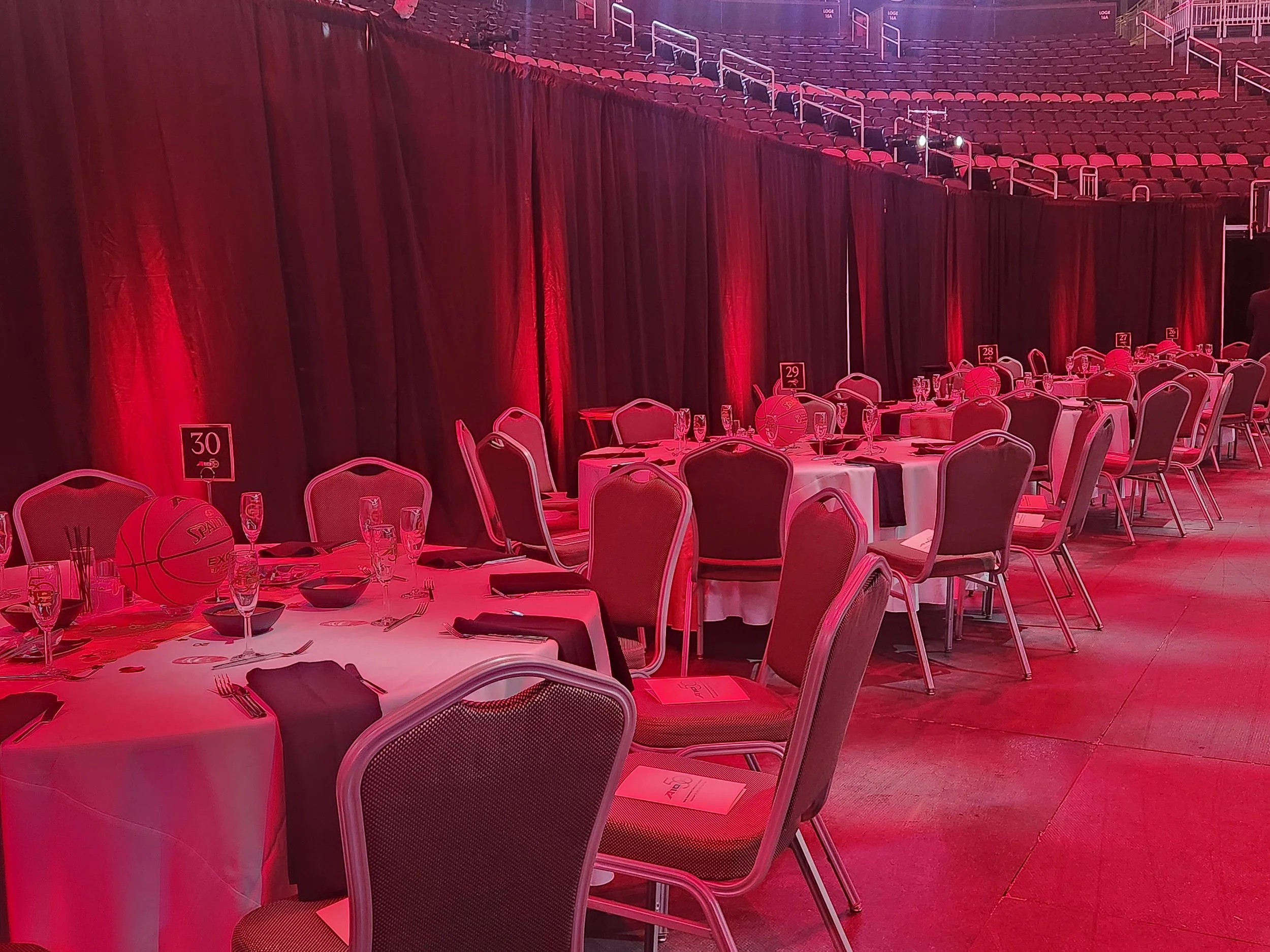 Round banquet tables set with glasses, plates, and black napkins, with a basketball decoration and table numbers, in a red-lit event space with chairs and stage curtains in the background. Up lighting Corporate event lighting solutions Gala Luncheon