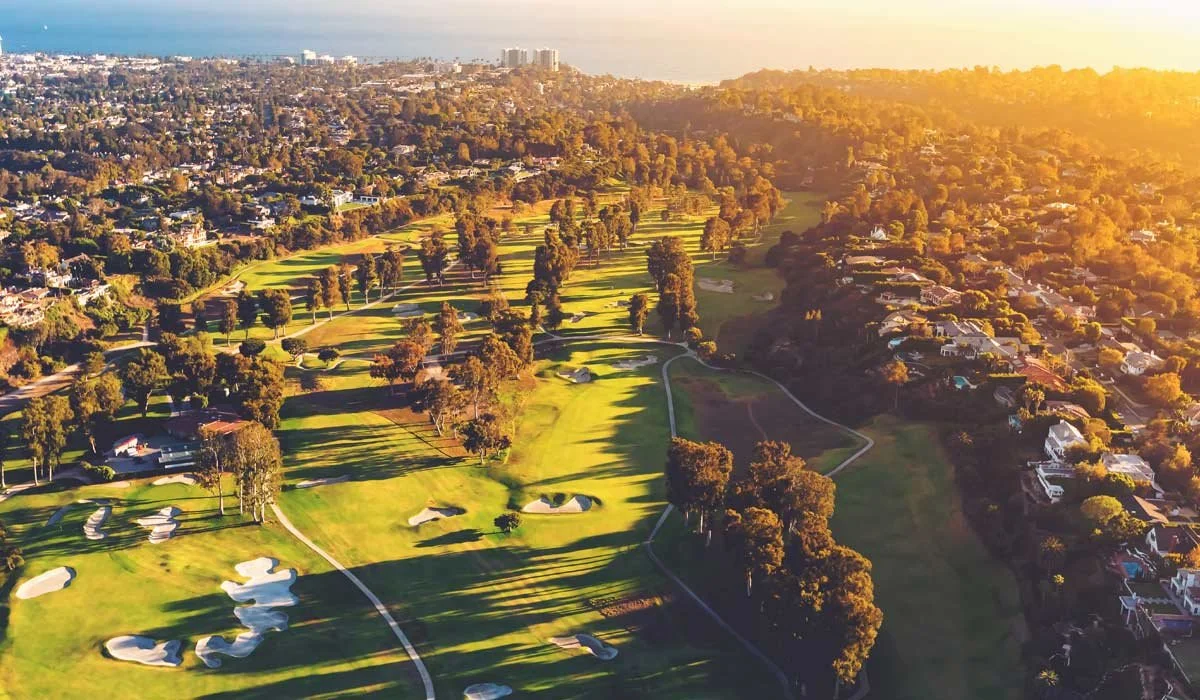 Aerial view of a golf course with sand traps and trees, surrounded by residential neighborhoods in a city, during sunset.