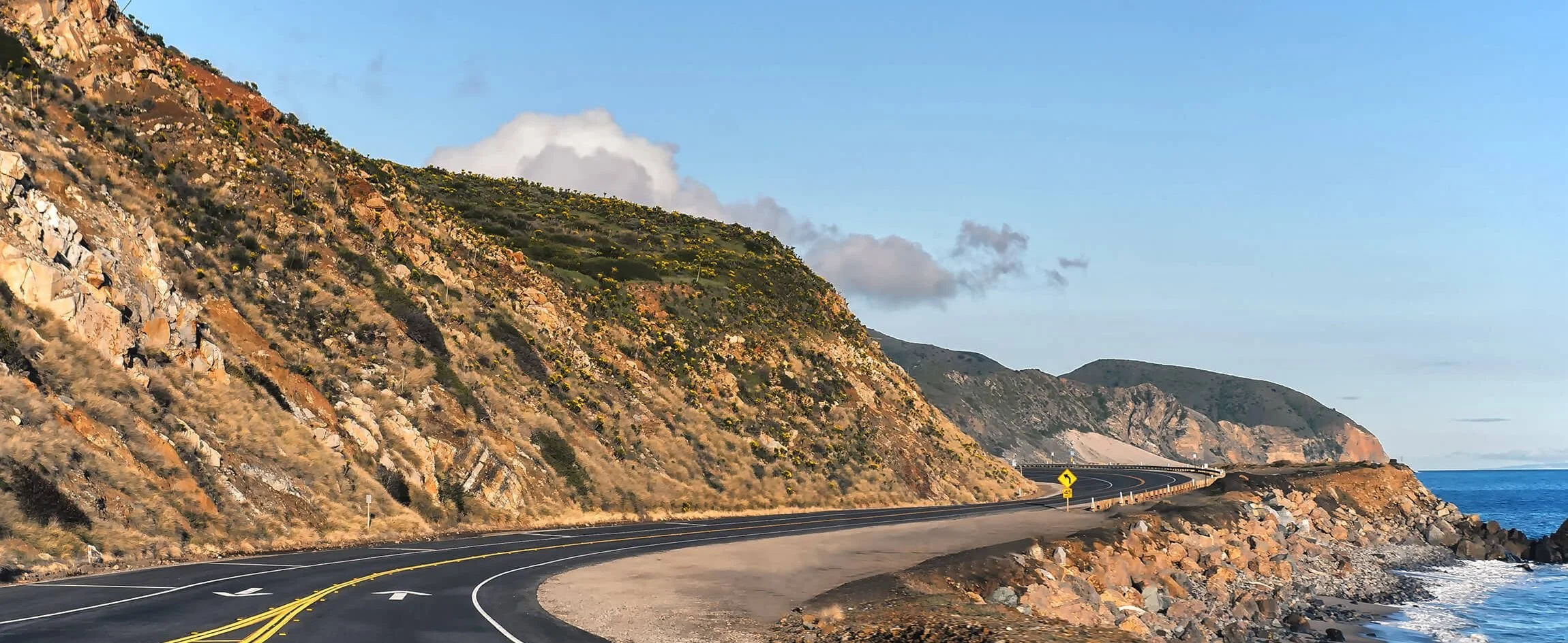 A winding coastal road runs along a hillside with rugged terrain, overlooking the ocean, under a partly cloudy sky.