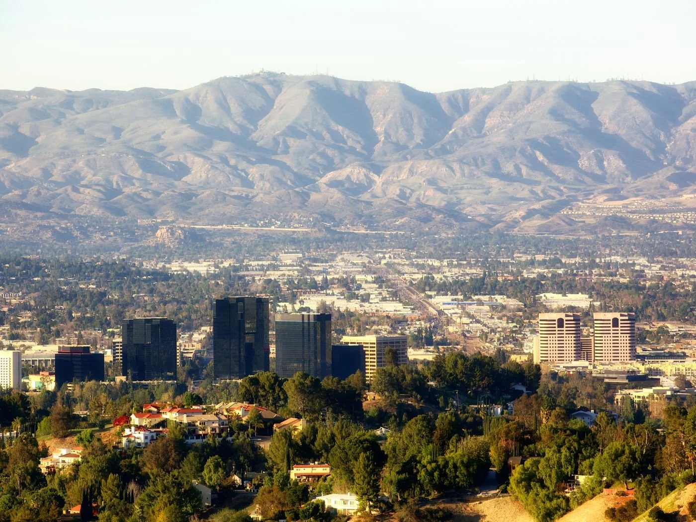 Cityscape with tall buildings in foreground, rolling hills and mountains in background under clear sky.