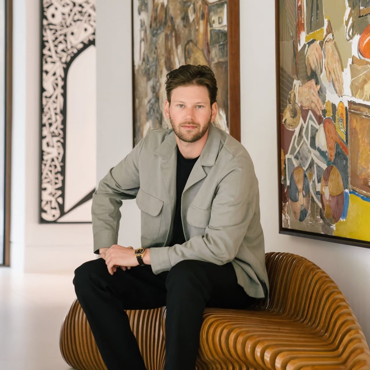 A man with dark hair and a beard sitting on a curved wooden bench in an art gallery, with colorful abstract paintings on the walls behind him.