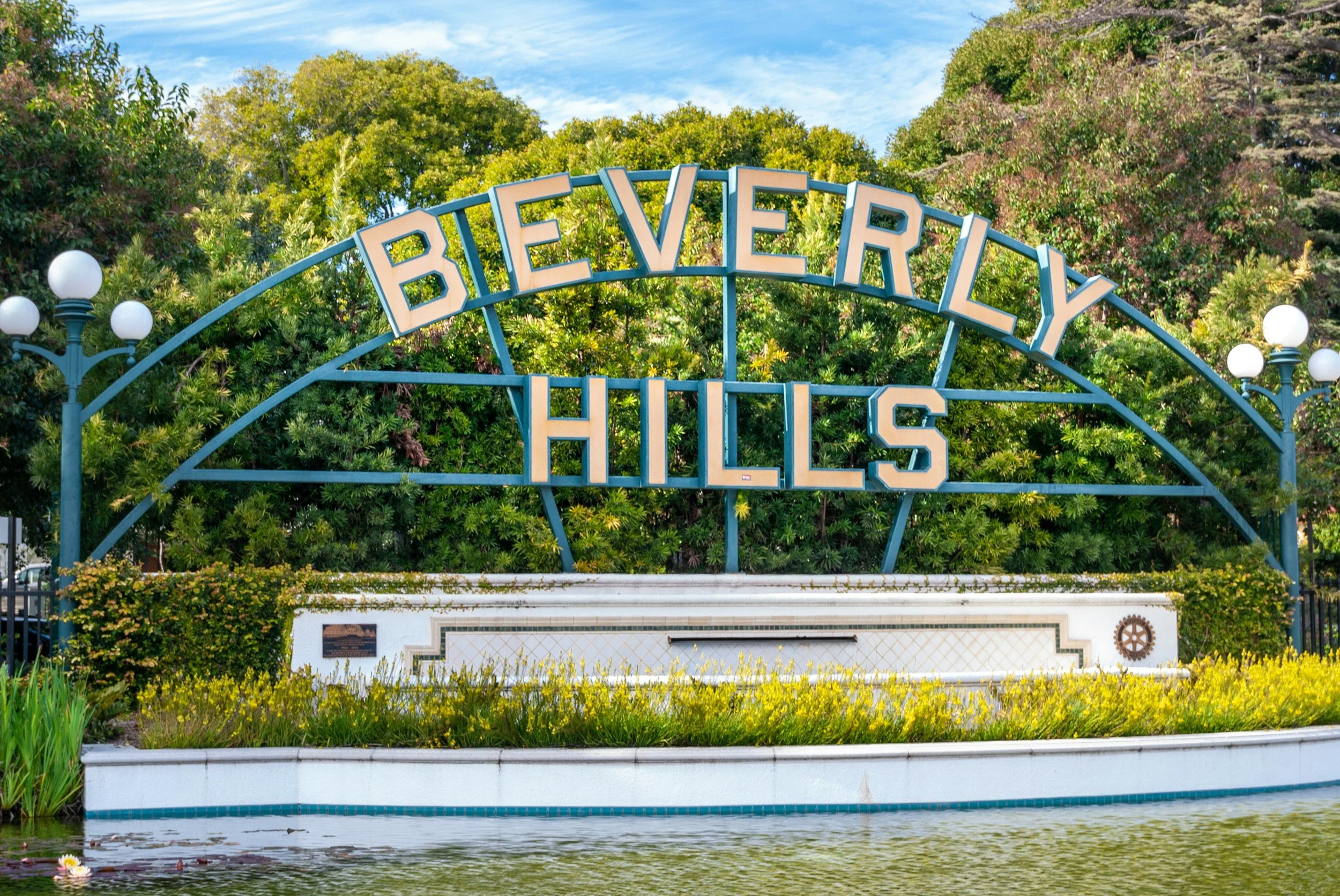 The Beverly Hills sign arch over a landscaped area with trees and a pond.
