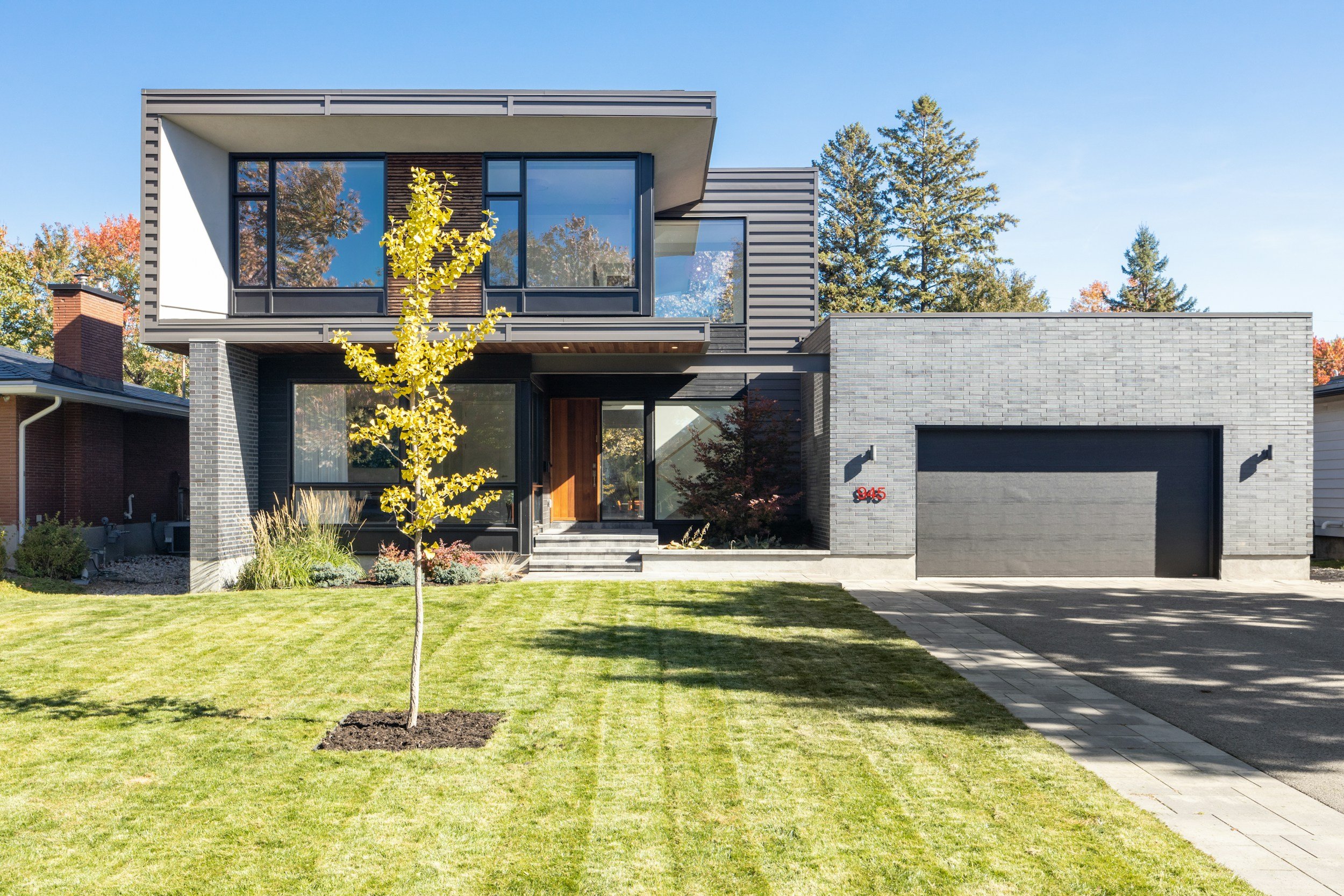 Modern two-story house with gray brick exterior, large glass windows, a small front yard with well-maintained grass, and a young tree, under a clear blue sky.