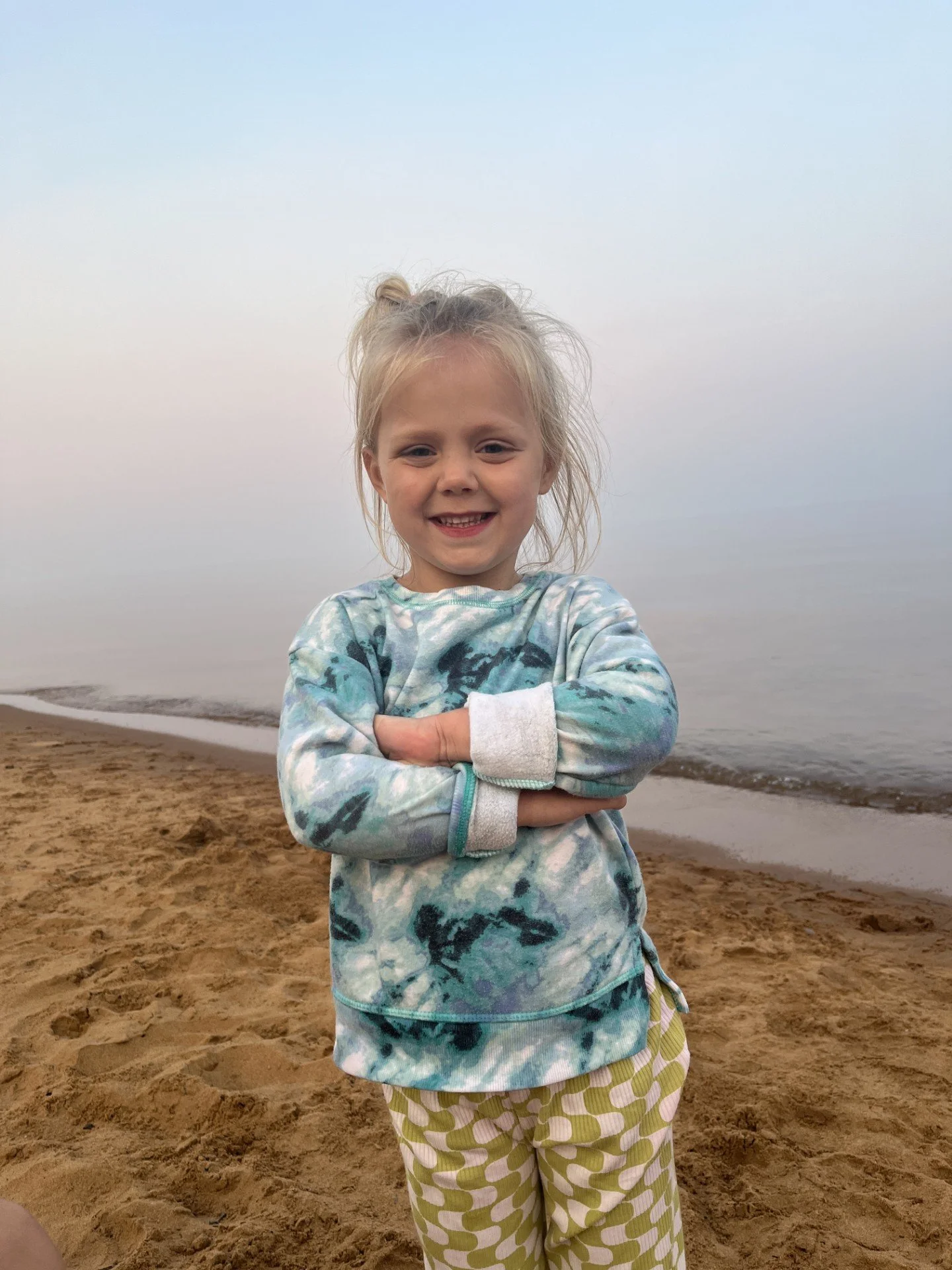 A smiling young girl with blonde hair standing on a sandy beach near the water, wearing a patterned hoodie and checkered pants.