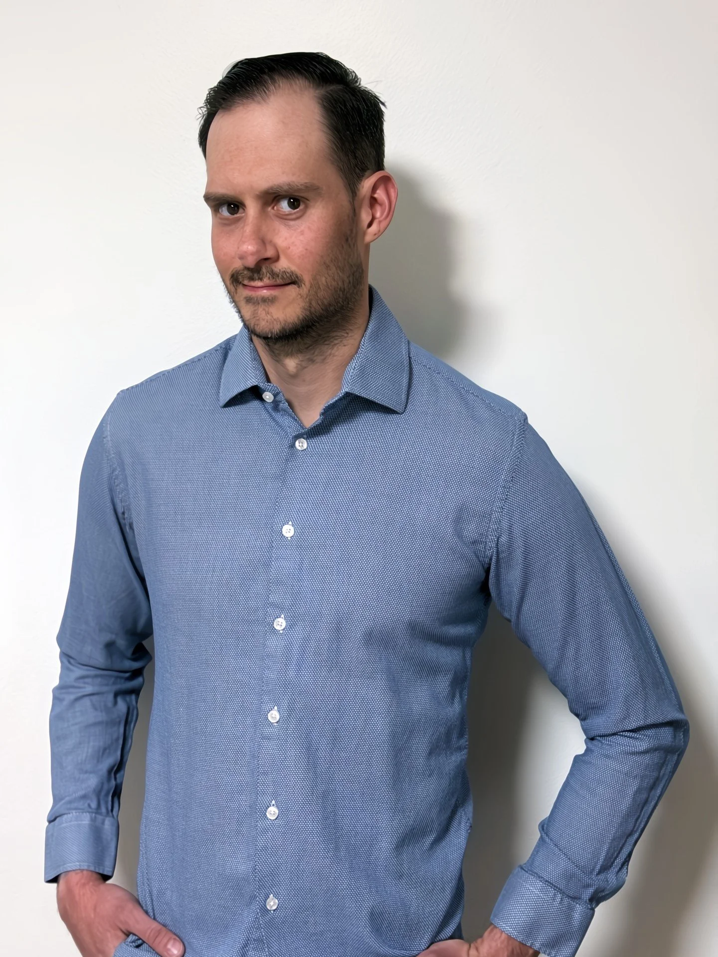Man in a blue button-up shirt standing against a white wall, with one hand on his hip, looking at the camera.