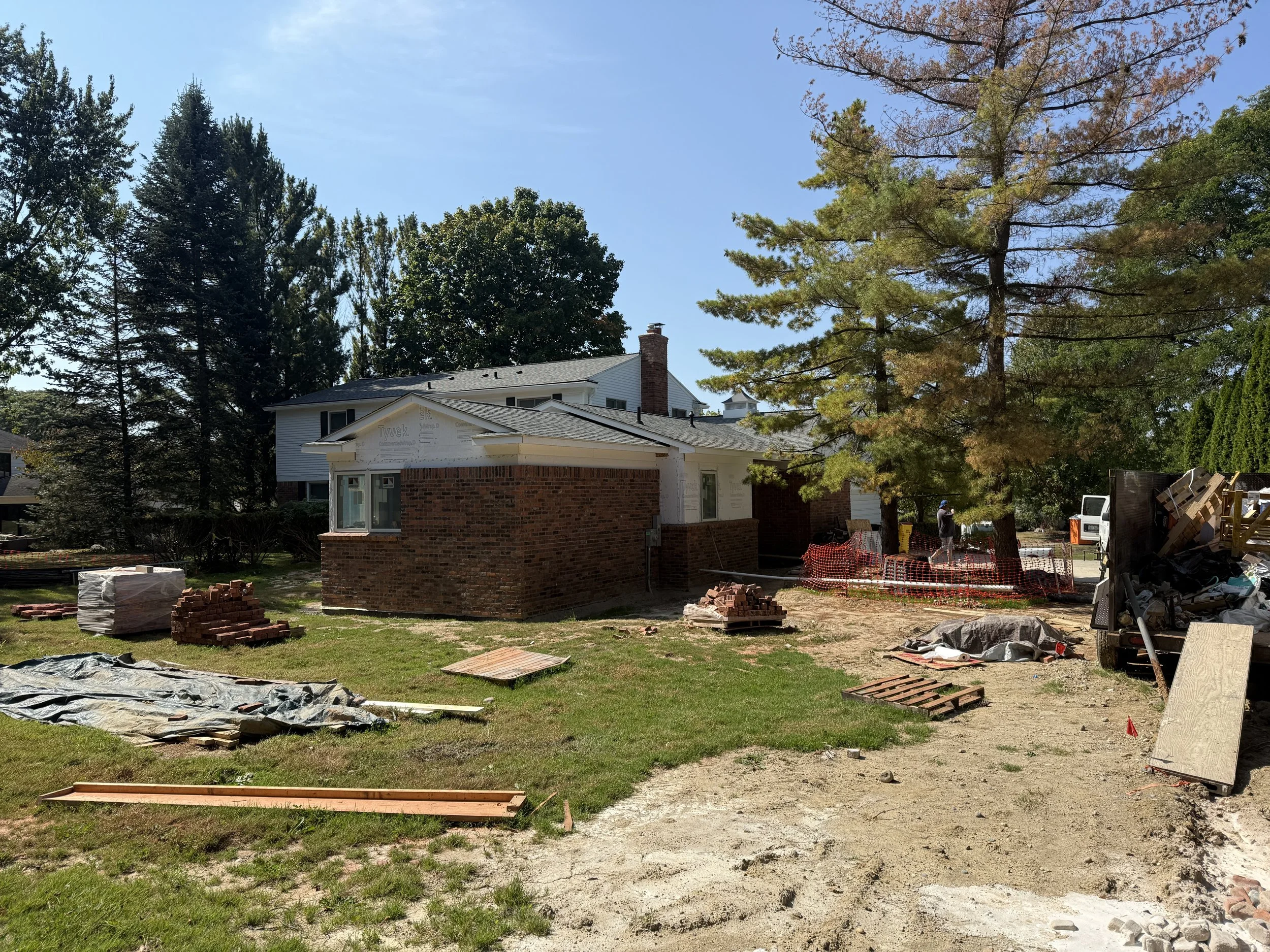 Stoneleigh Residence construction progress showing exterior brickwork underway on the new addition, surrounded by mature pines and construction materials — designed by KJF Studio in Bloomfield Township, Michigan.